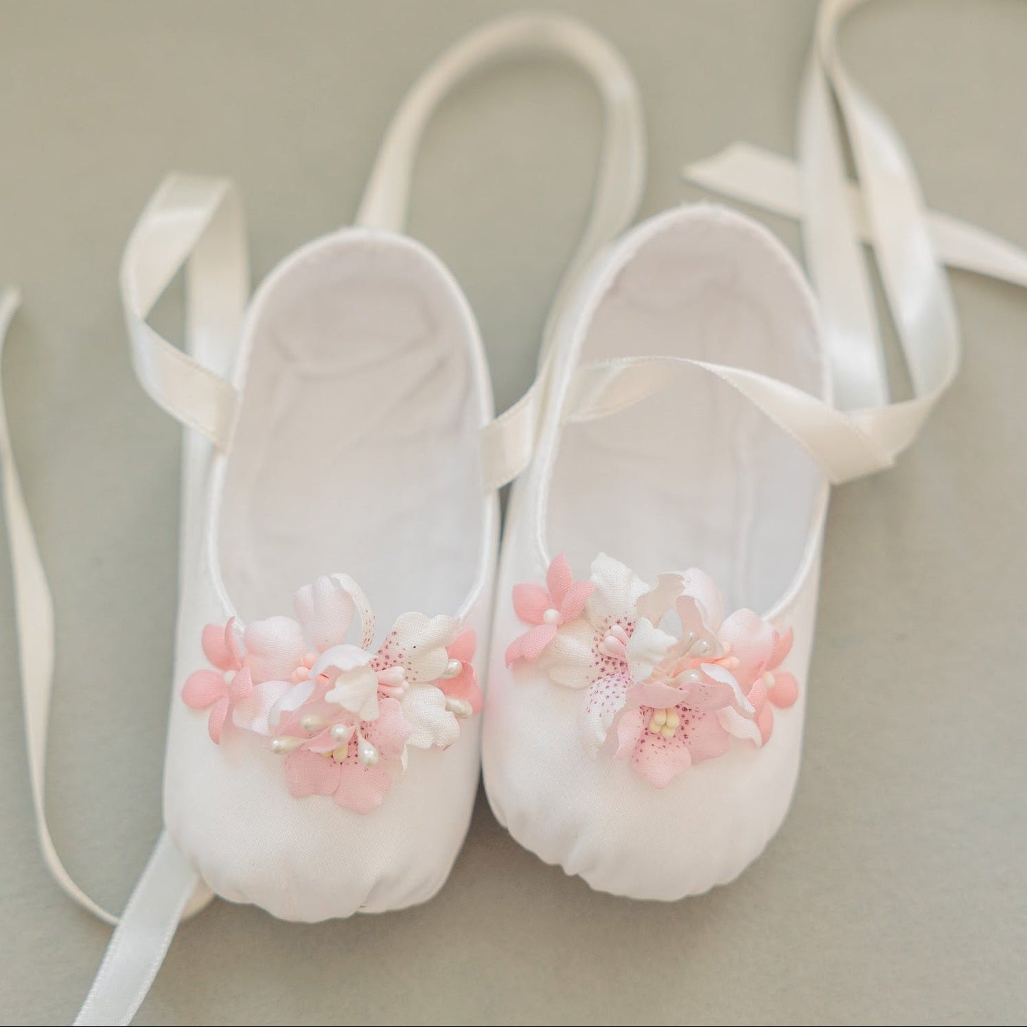 A pair of white ballet pointe shoes decorated with pink and white fabric flowers, shown on a neutral gray background.