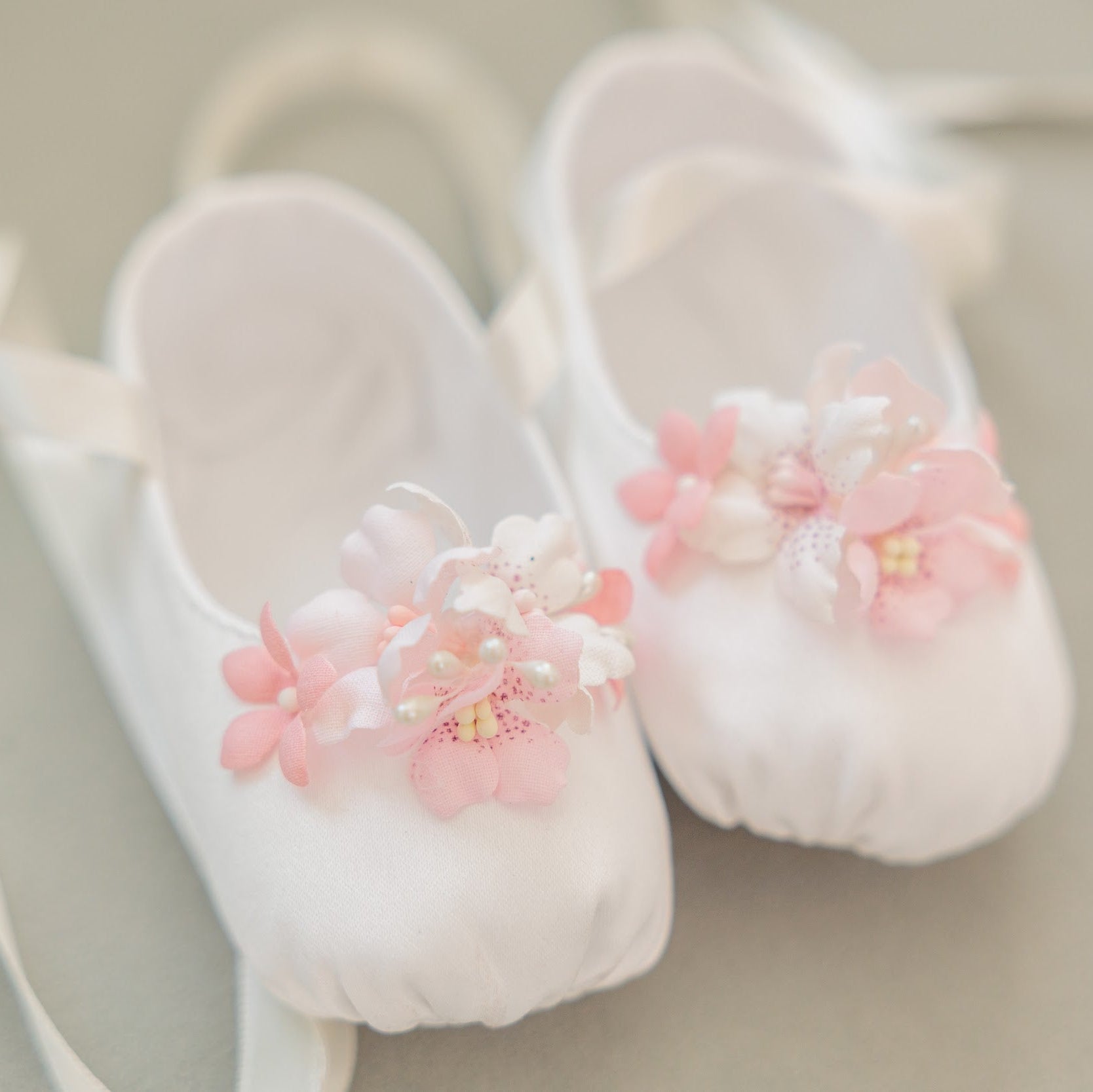 A pair of white satin ballet pointe shoes decorated with pink and white fabric flowers, resting on a neutral beige background.