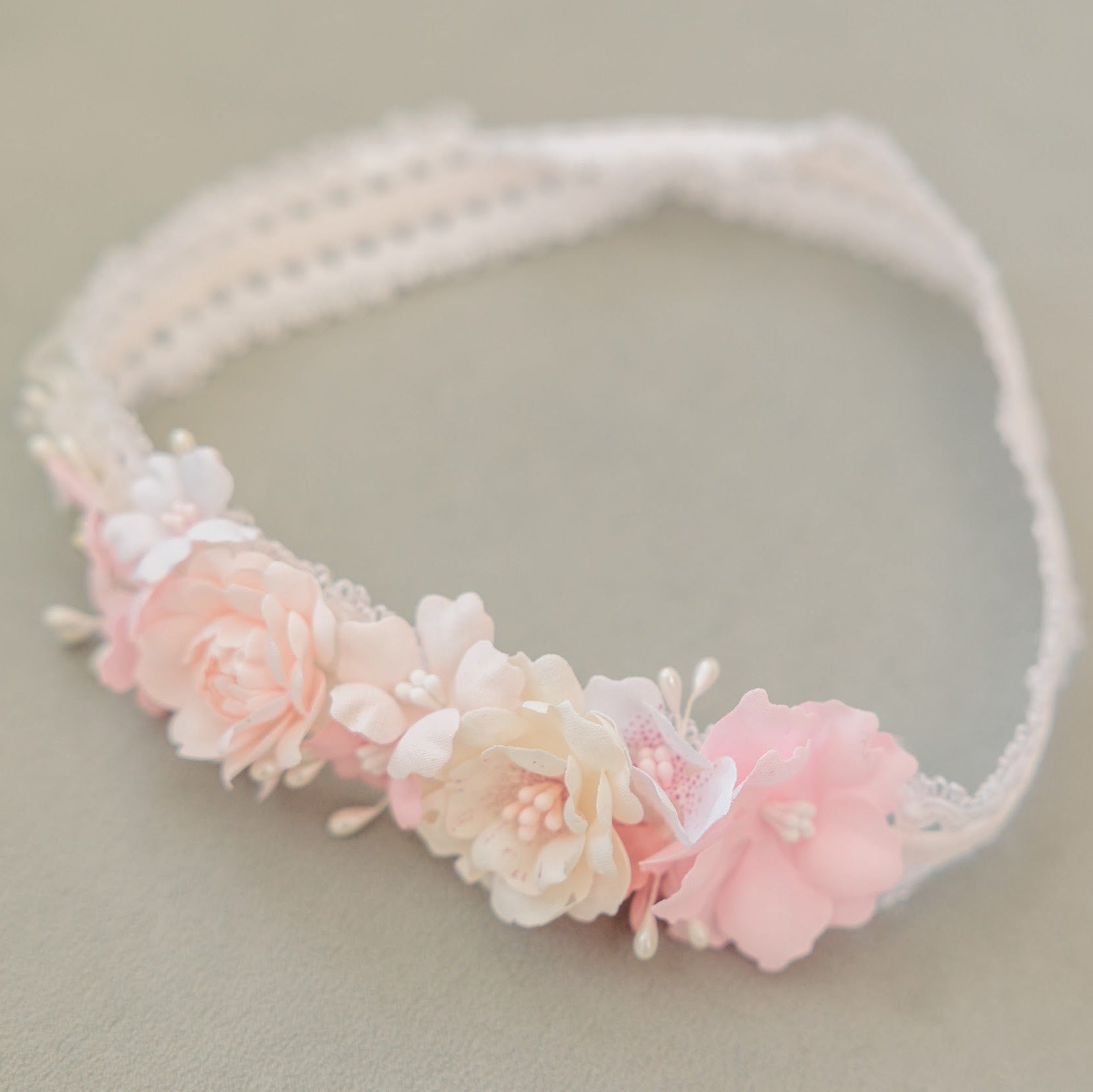 A delicate white lace headband decorated with a row of soft pink and cream fabric flowers, photographed on a neutral beige background.