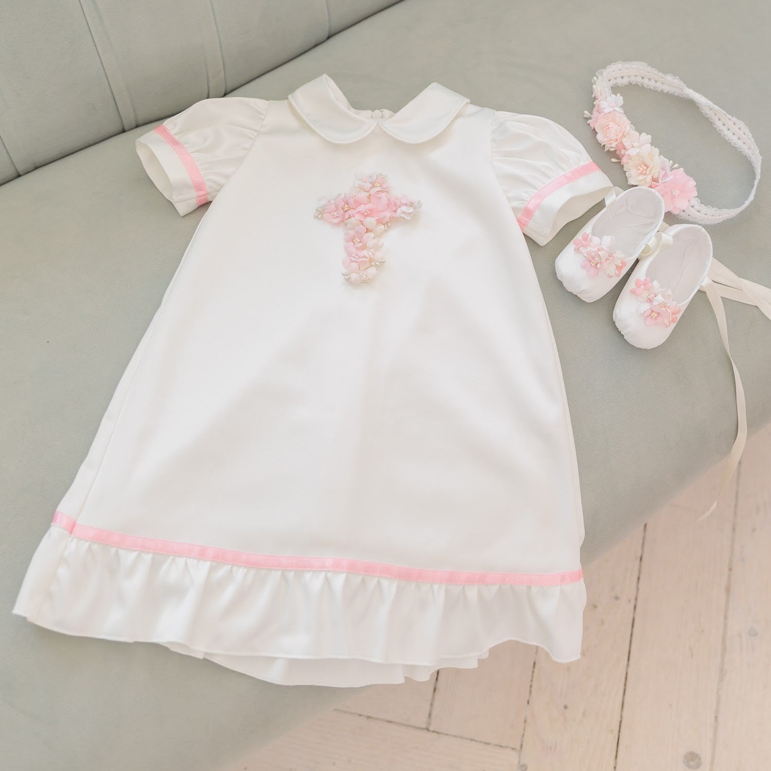 A baby girl's white dress outfit set with pink trim, featuring a short-sleeve dress, matching booties, a floral headband, and a small bow, laid out on a gray tufted sofa.