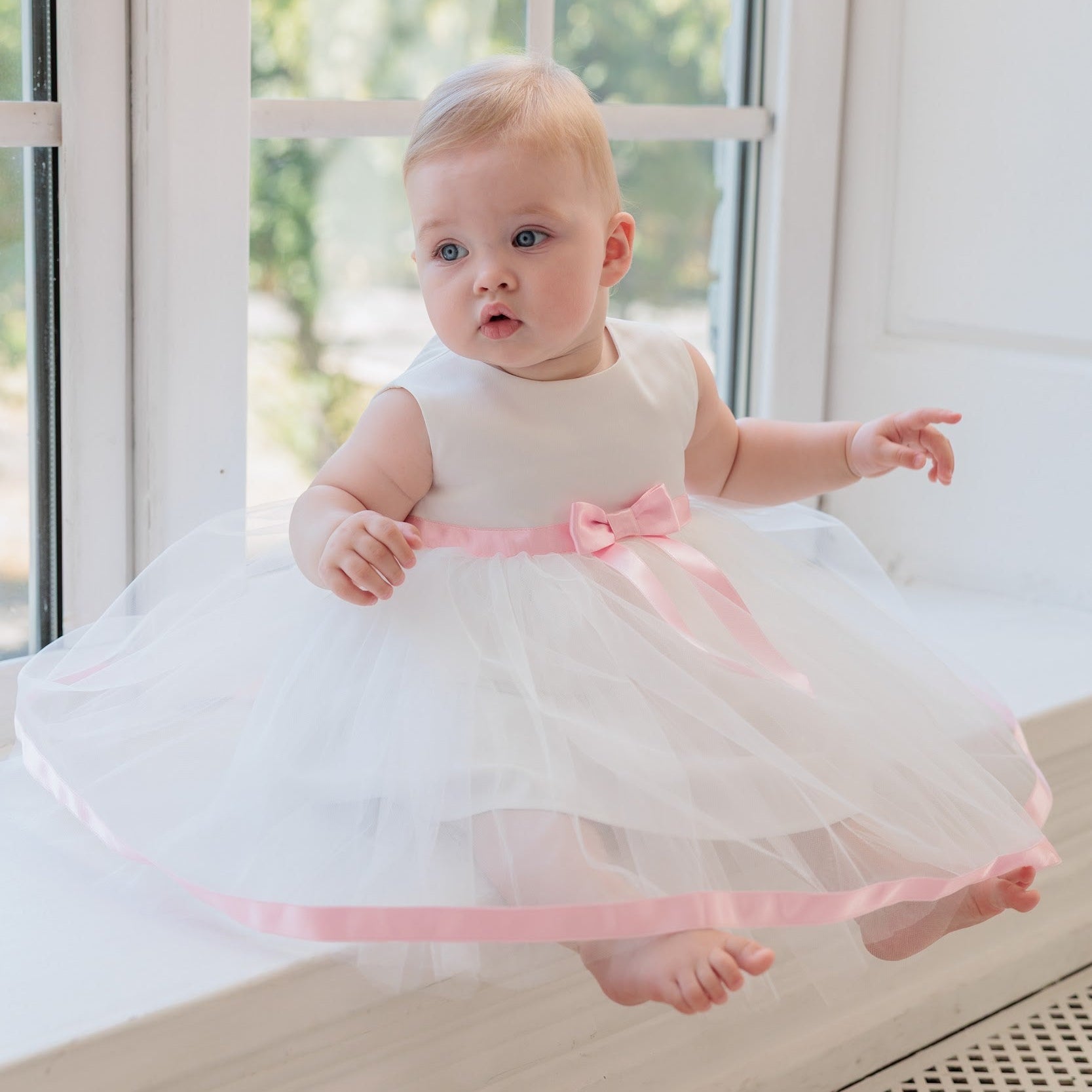 A baby girl wearing a white sleeveless tulle dress with a pink ribbon bow and trim, sitting on a white windowsill.