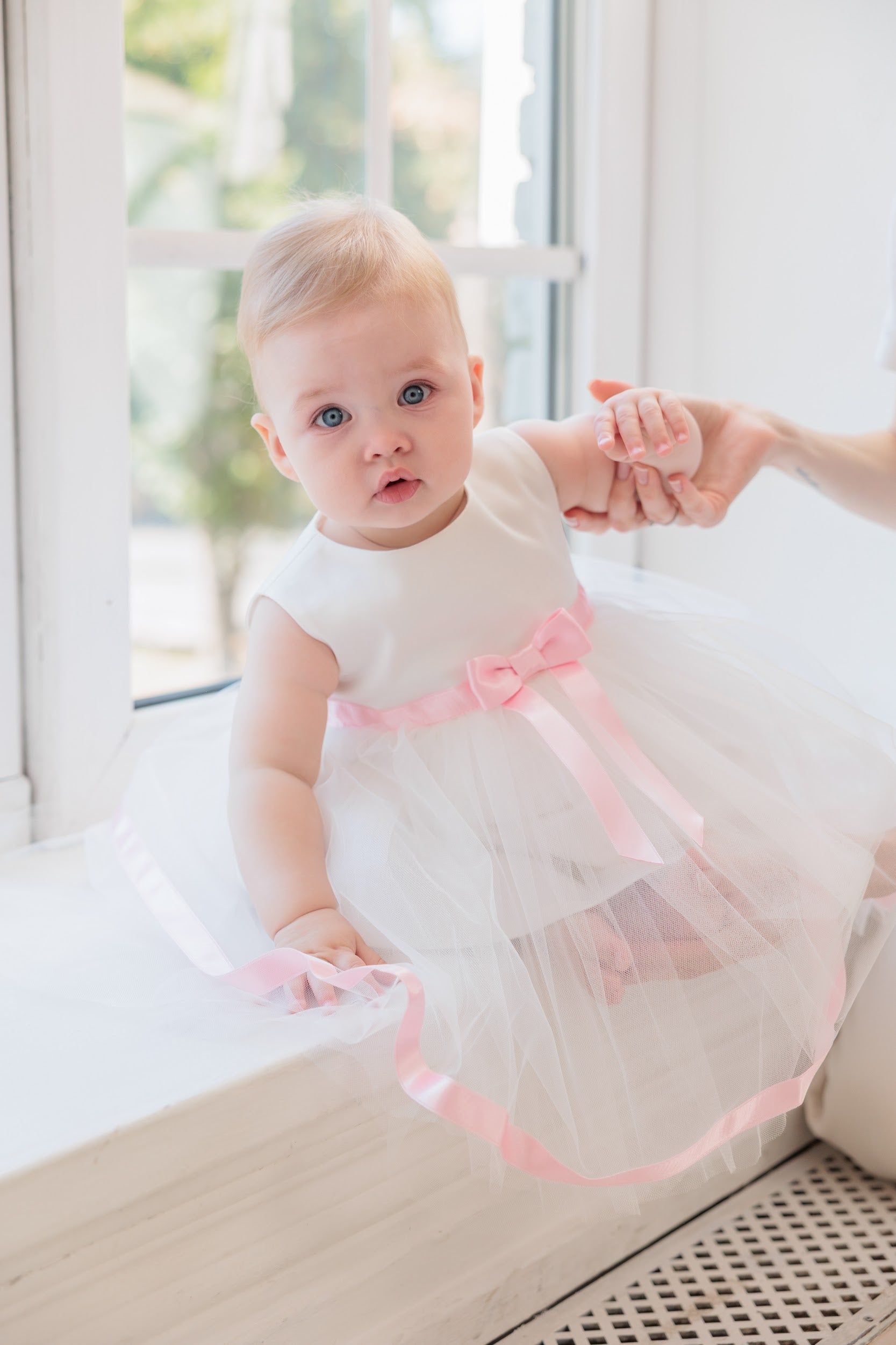 A baby girl wearing a white sleeveless tulle dress with a pink ribbon bow and trim, sitting on a windowsill while being held up by an adult's hand.
