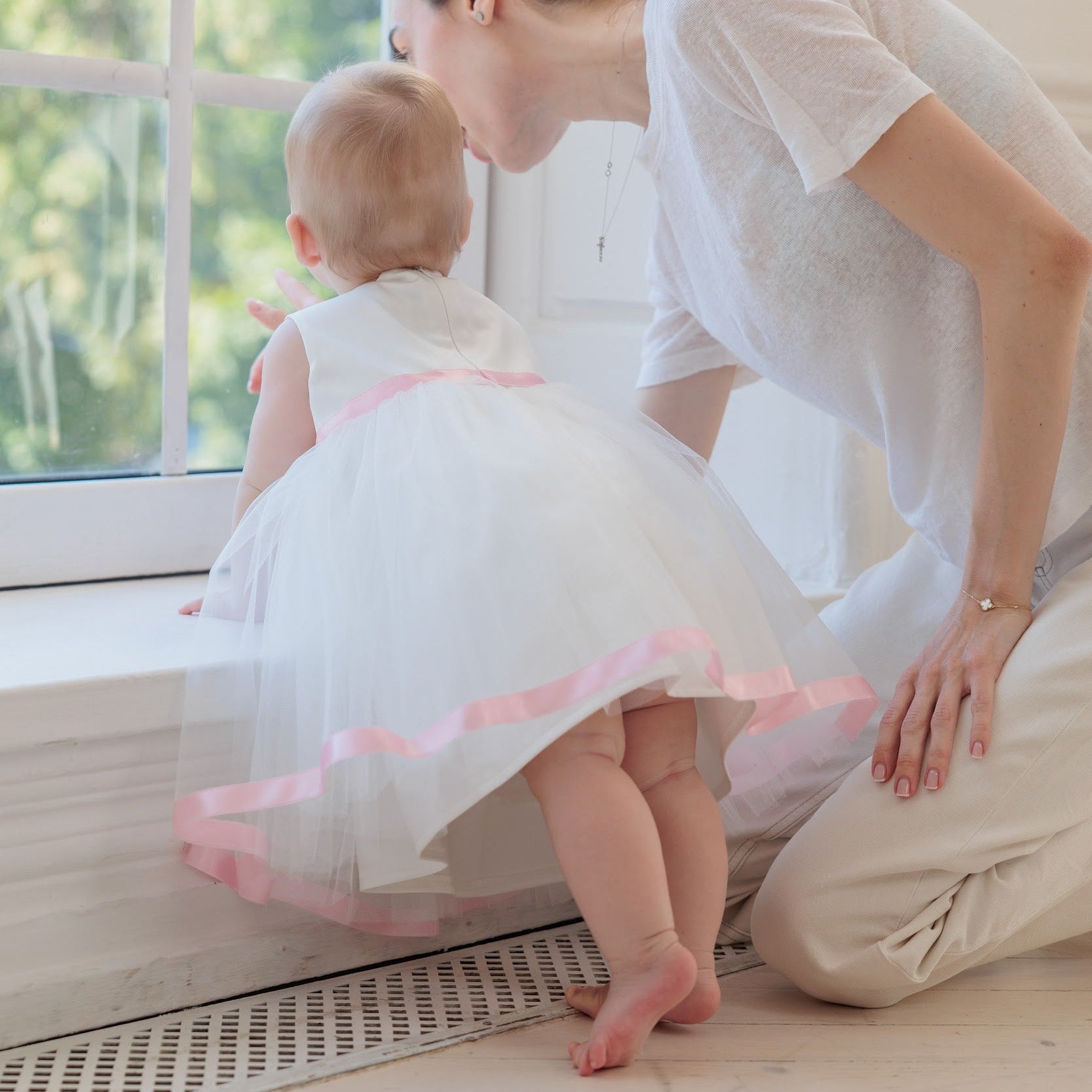 A woman kneels beside a baby in a white tutu dress with pink trim, looking at the child by a bright window in a light, airy room.
