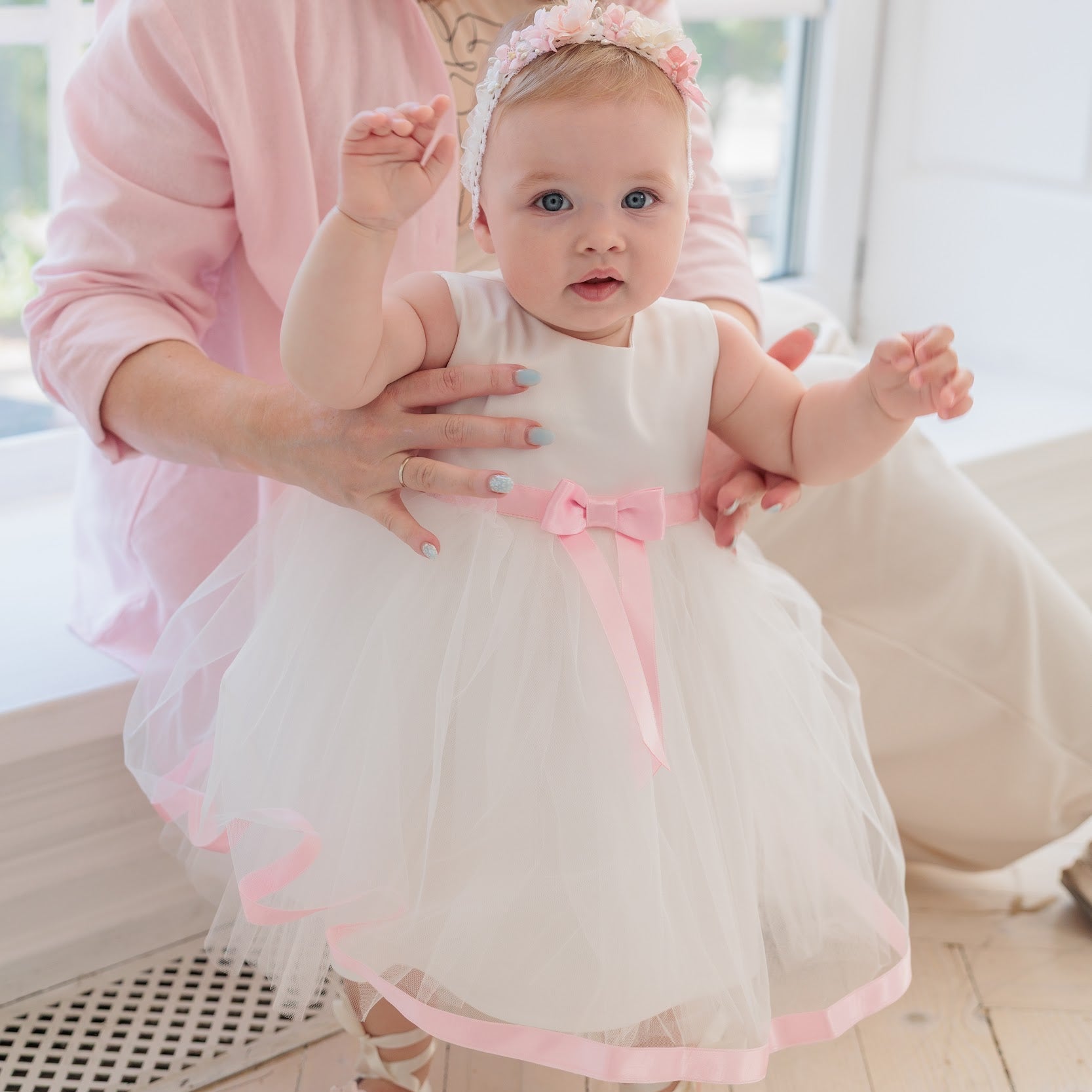 A baby girl in a white tulle flower girl dress with a pink ribbon and bow, wearing a pink floral headband, being supported by a woman in a pink shirt while walking indoors.