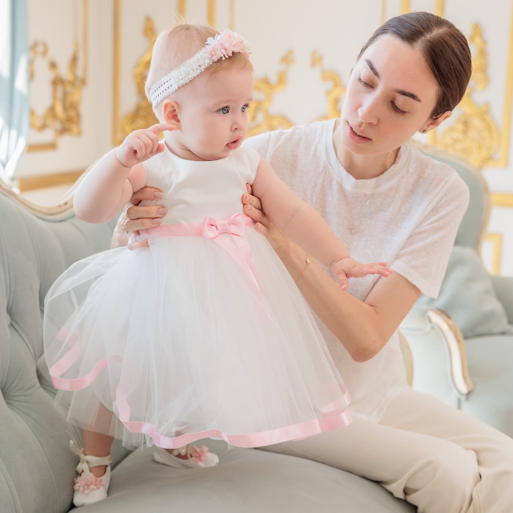 A woman sits on a light gray tufted sofa holding a toddler wearing a white tutu dress with a pink ribbon and bow, in an elegant room with gold-trimmed walls.