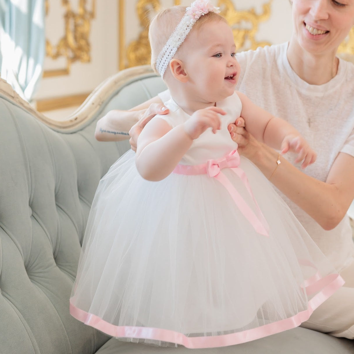 A woman sits on an ornate light blue-green sofa in a luxurious room, adjusting a baby girl's white tulle first birthday dress with a pink ribbon and bow. The baby also wears a white headband with a bow.