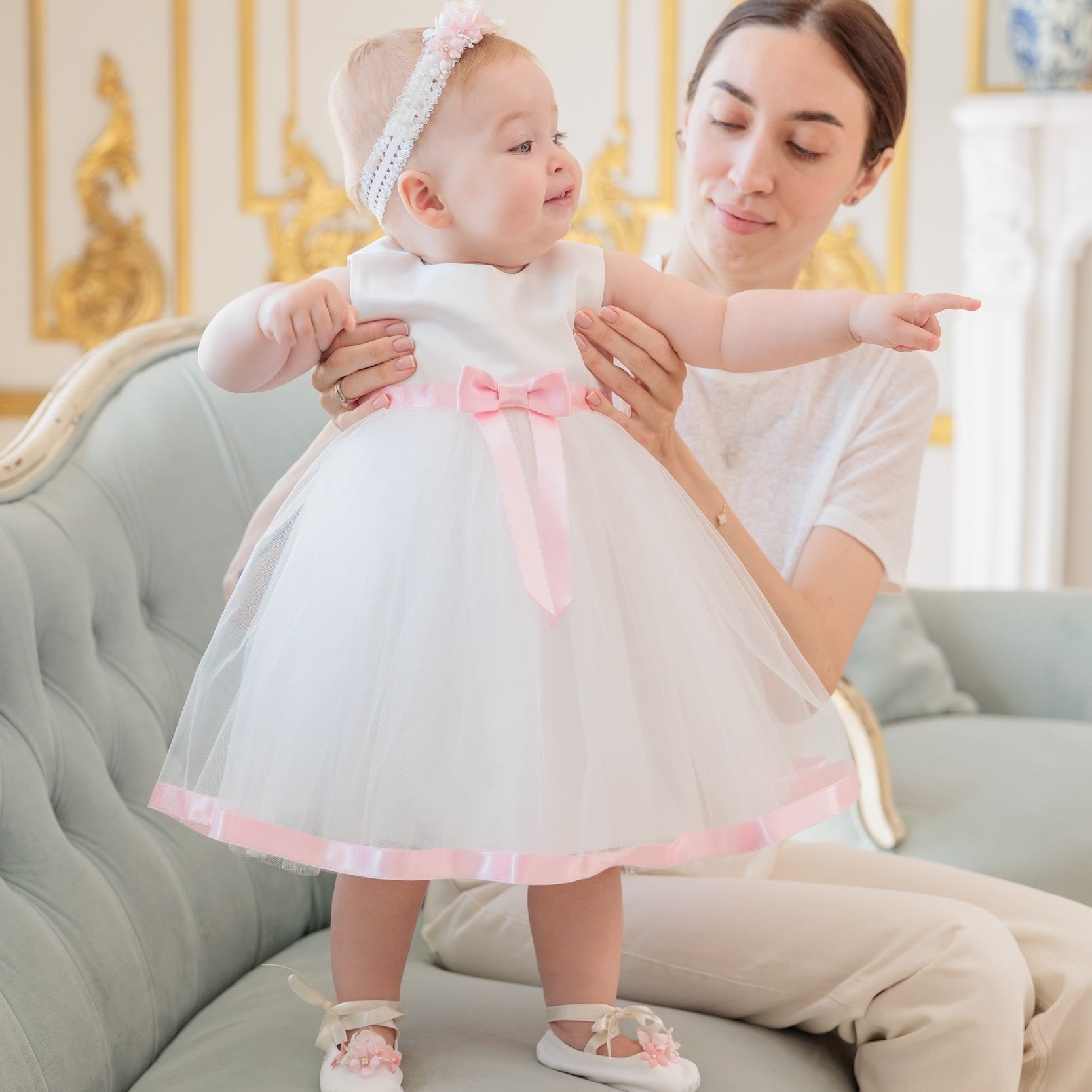 A baby girl in a white tulle first birthday dress with a pink bow and headband stands on her mother's lap, pointing to the side, set in a classic elegant room with a tufted sofa.