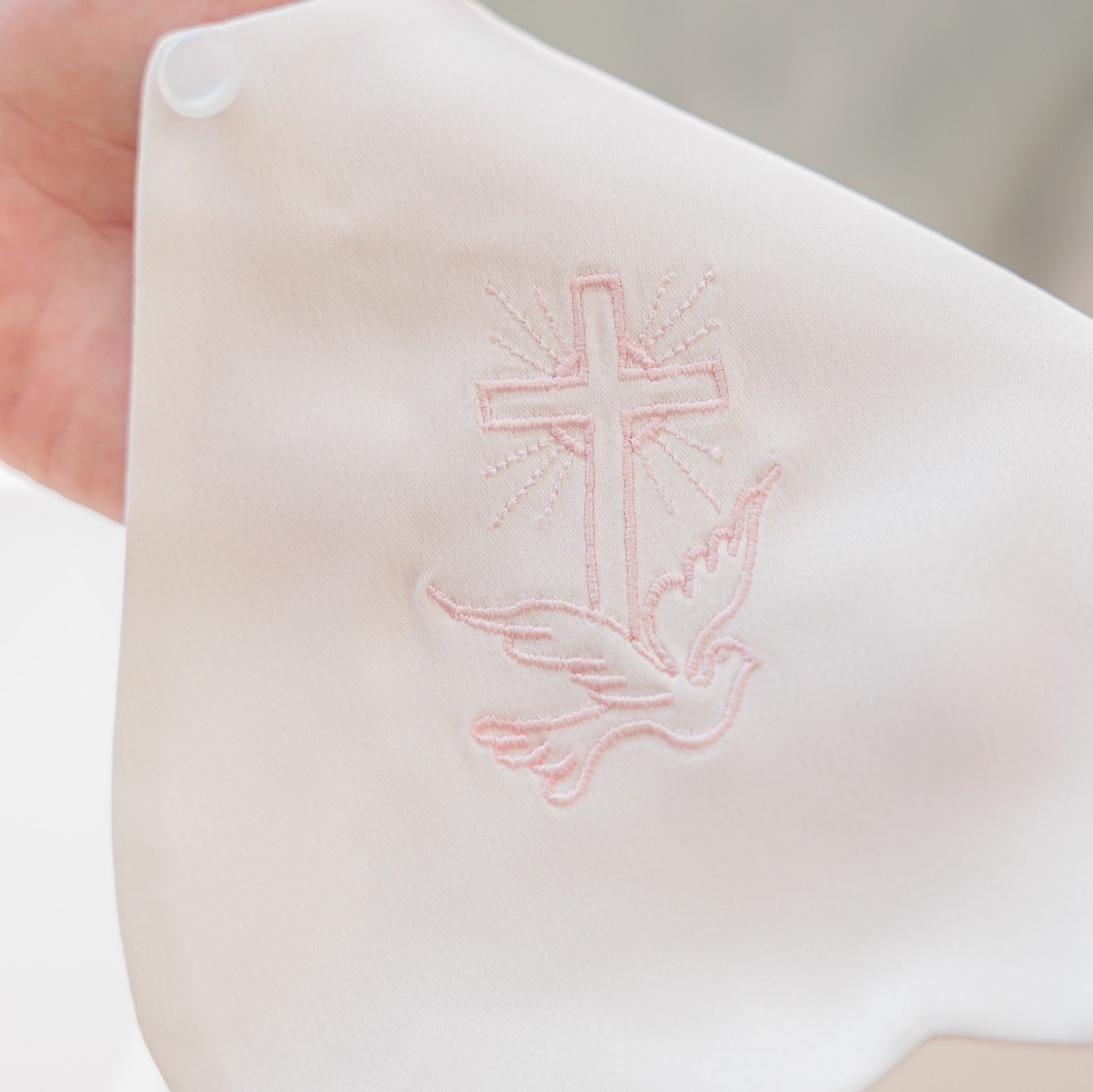 A close-up of a white or off-white fabric item being held by a hand, featuring a light pink embroidered cross and dove design on the front.