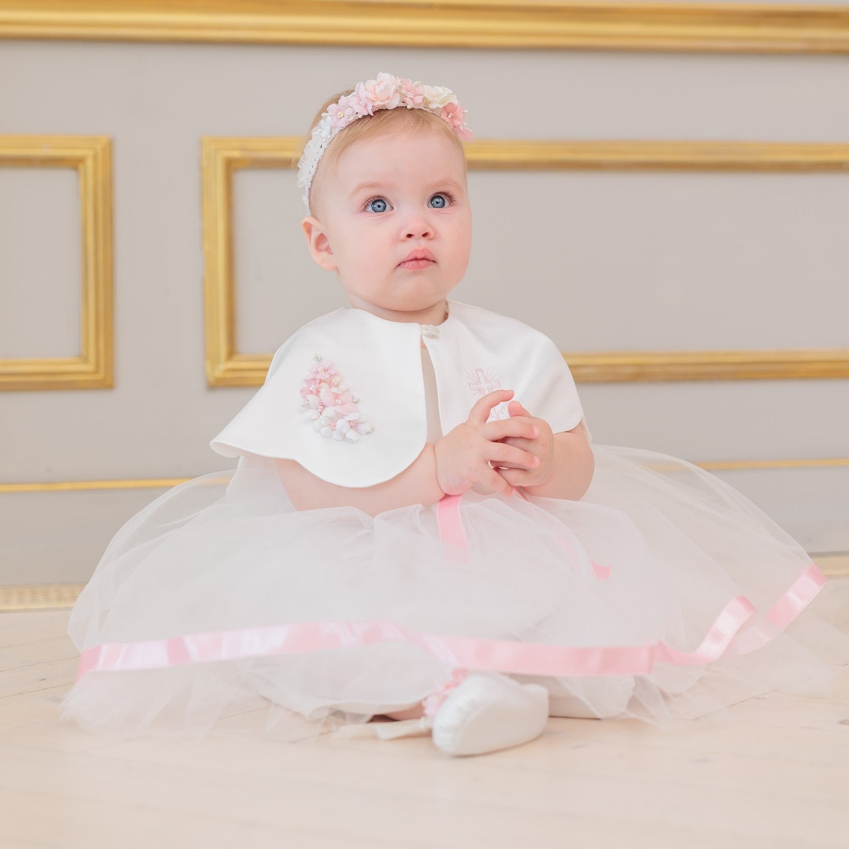 A baby girl wearing a white and pink first birthday tutu dress with a matching headband, sitting on a light wooden floor in front of a beige wall with gold picture frames.