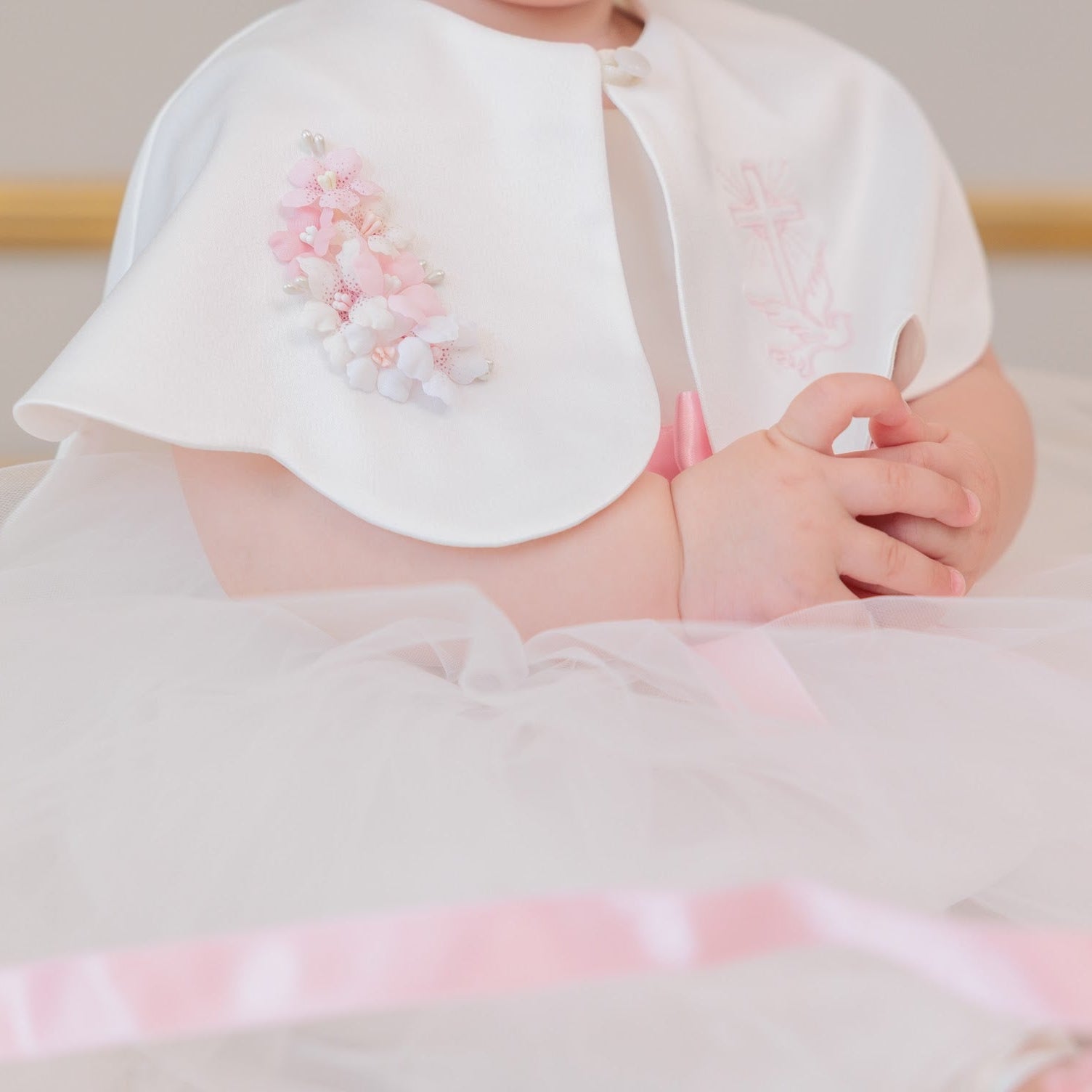 A close-up of a baby wearing a white tulle dress with a matching white bonnet decorated with pink and white floral embroidery, sitting in a soft indoor setting.
