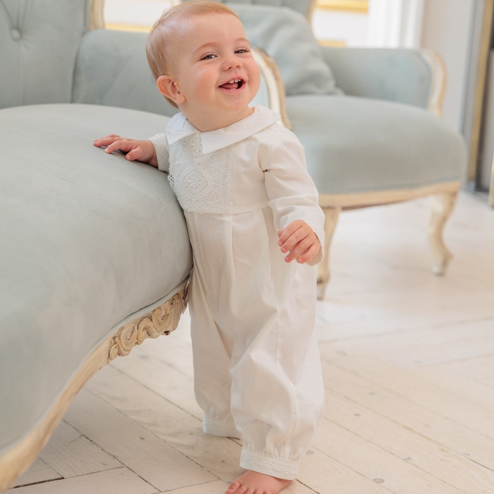 A baby standing on a wooden floor and leaning on a light blue velvet chaise lounge, wearing a white long-sleeve baby jumpsuit with a lace collar.