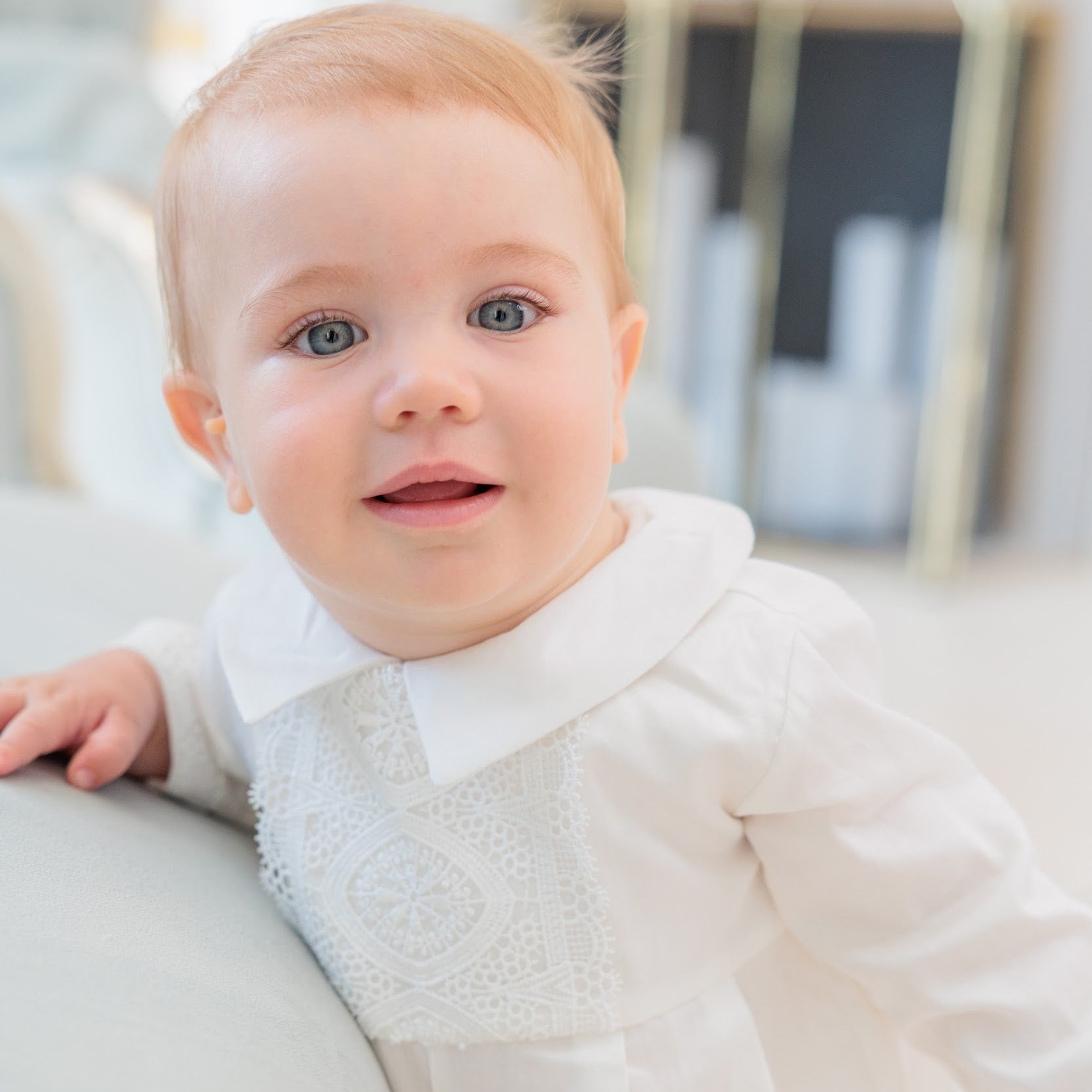 A baby girl with light hair and blue eyes wearing a white long-sleeve dress with a white collar and embroidered yoke, leaning on a light-colored sofa in a bright, modern room.