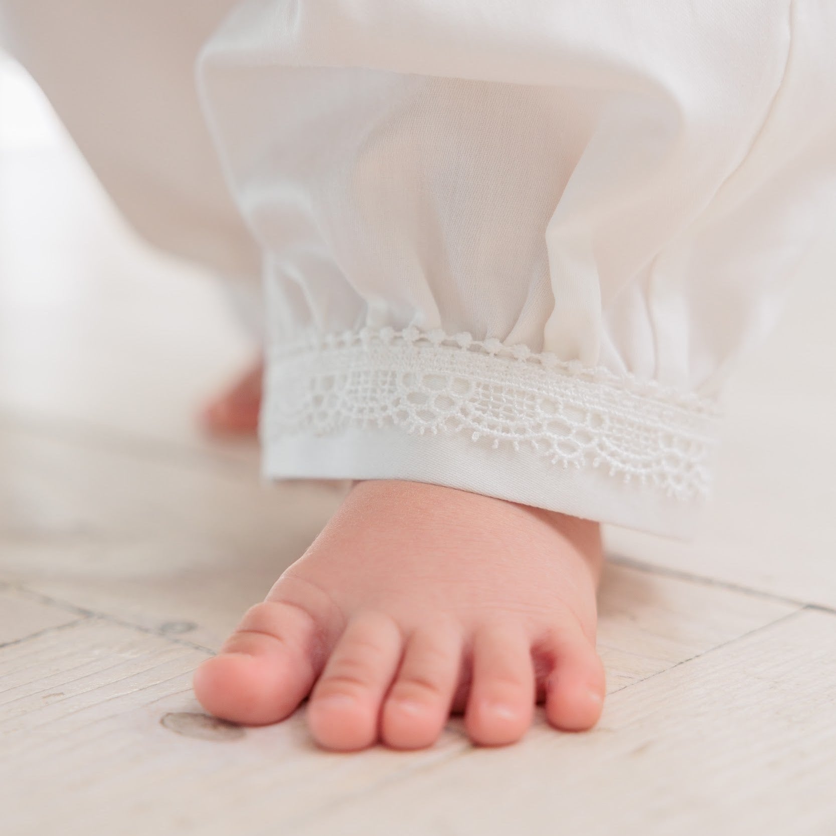A close-up of a baby's bare foot stepping onto a light-colored wooden floor, wearing a white long-sleeve dress with delicate lace trim at the ankle.