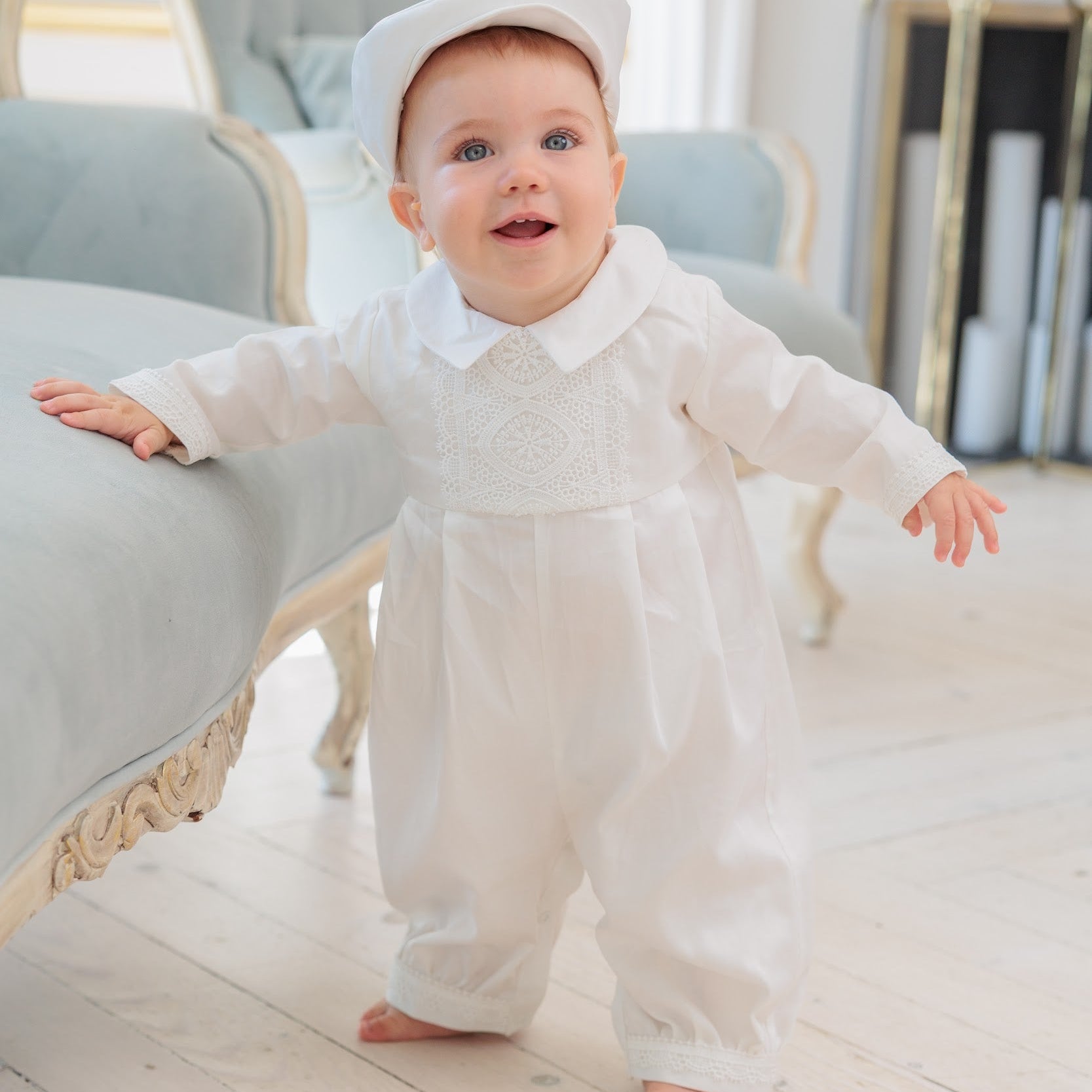 A baby wearing a white long-sleeve baby romper with a matching white baby hat, standing on a light wooden floor in a bright, elegant room.