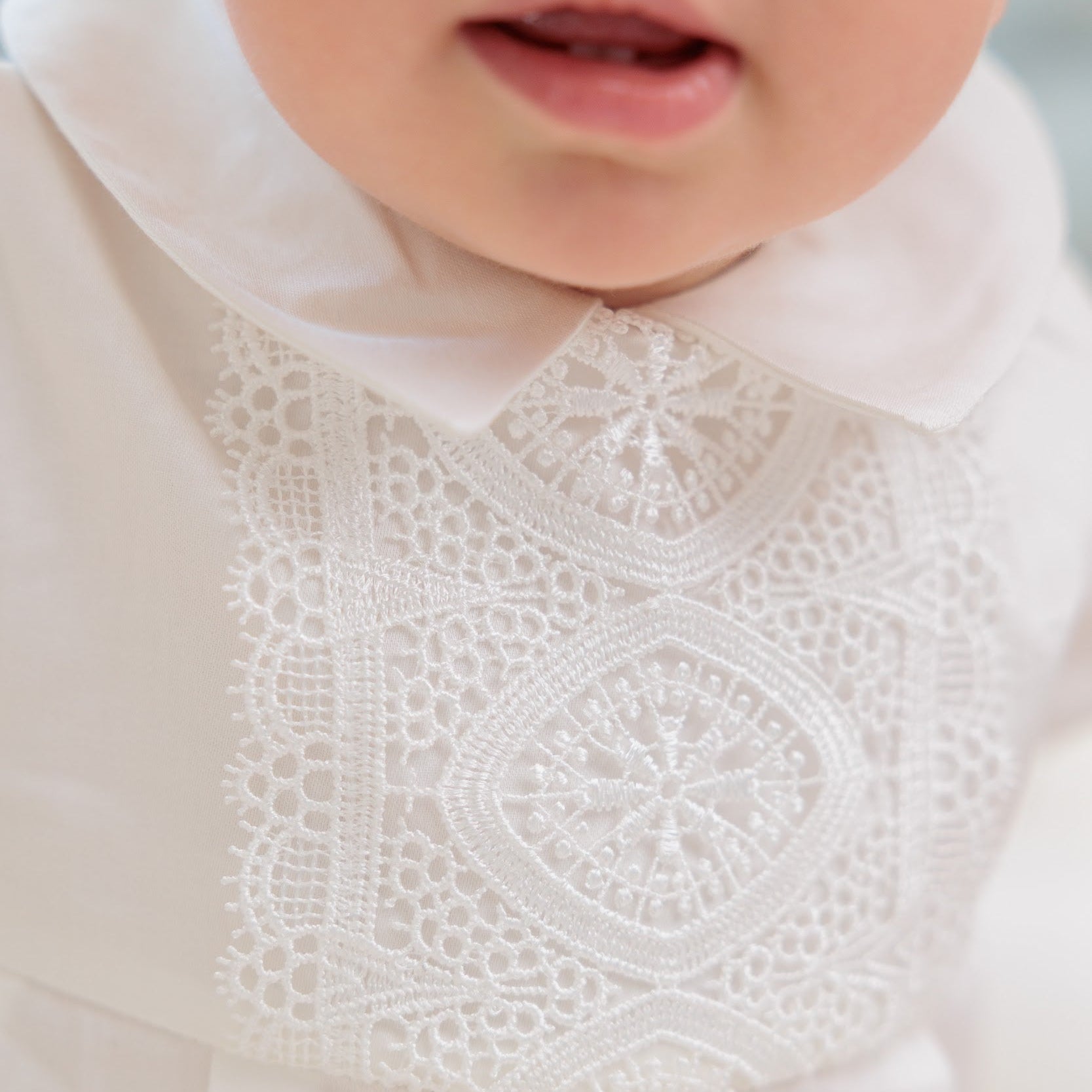 Close-up of a baby wearing a white lace baby dress with a Peter Pan collar and intricate embroidered lace panel on the chest.