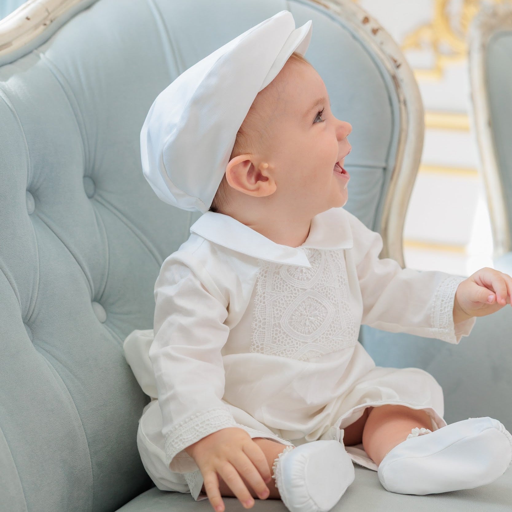 A baby wearing a white long-sleeve romper, matching white hat, and white booties sits on a light blue tufted armchair in an elegant room.