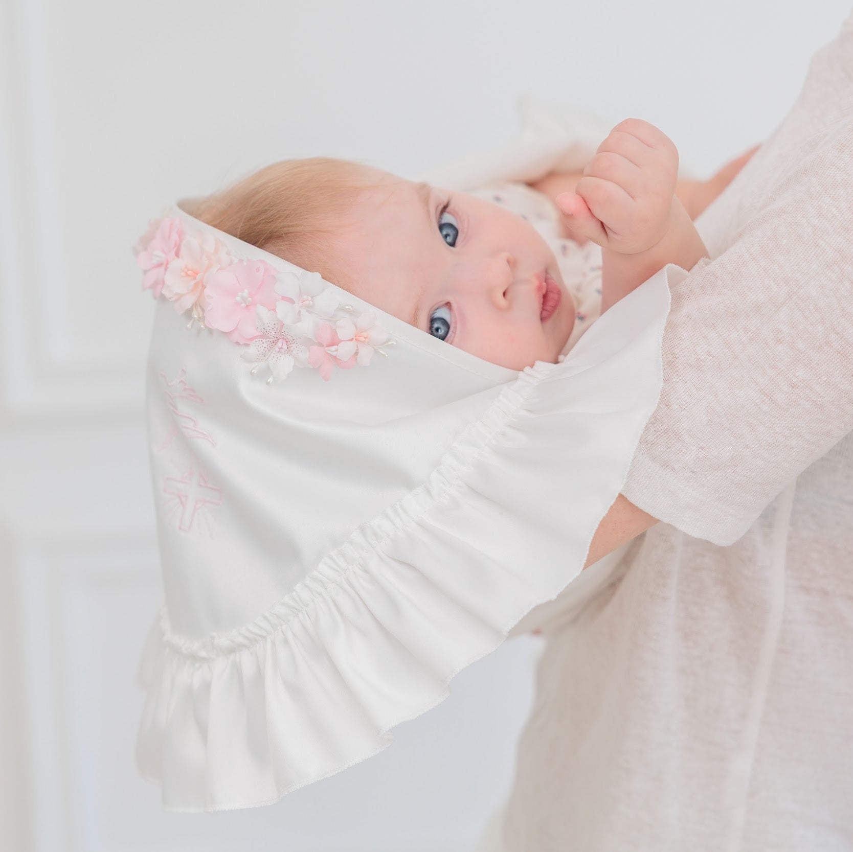 A baby wearing a white bonnet with pink floral embroidery and a ruffled edge, held by an adult in a soft white top against a bright white background.