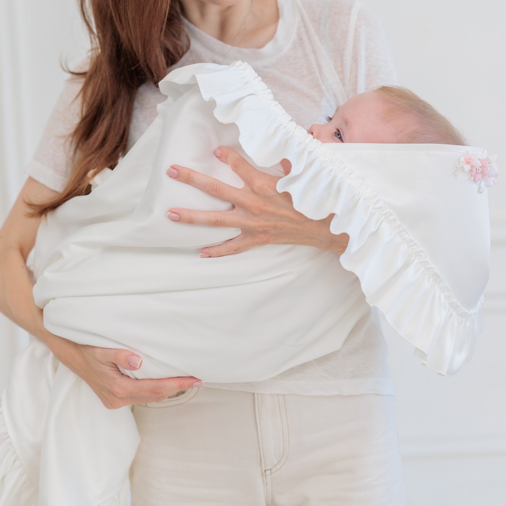 A woman cradles a baby wrapped in a soft white ruffled swaddle blanket, set against a bright, minimalist white paneled background.