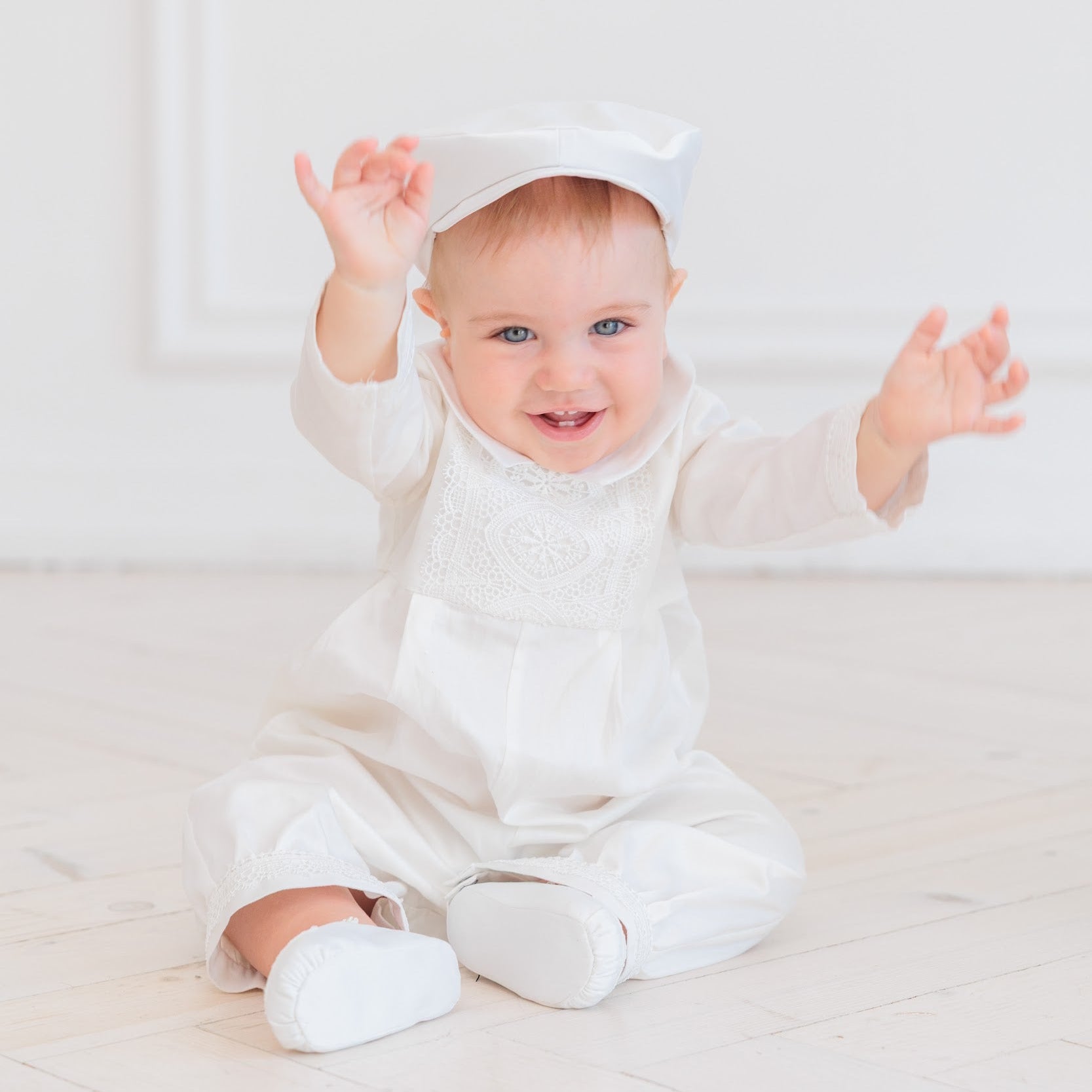 A baby sitting on a light wooden floor wearing a white long-sleeve romper with lace detailing, a matching white bonnet, and white booties.