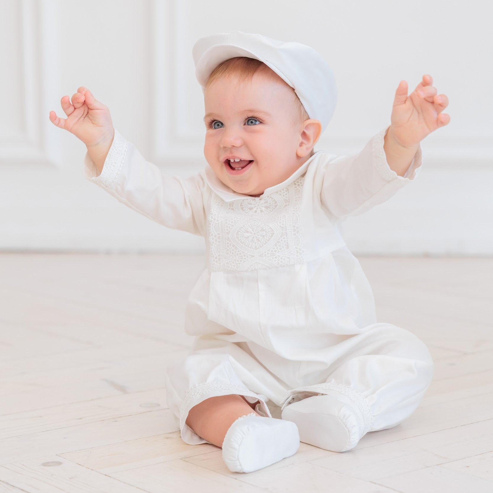 A smiling baby sitting on a light wooden floor wearing a white long-sleeve romper, matching bonnet, and white booties, with arms raised in the air.