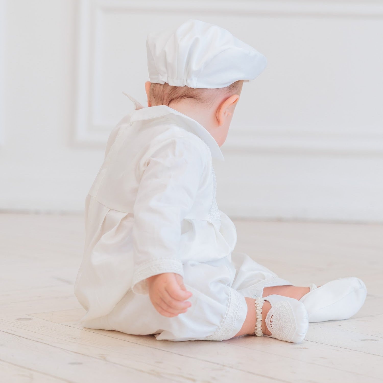 A baby sitting on a light wooden floor, viewed from behind, wearing a white long-sleeve romper, matching white bonnet, and white socks.