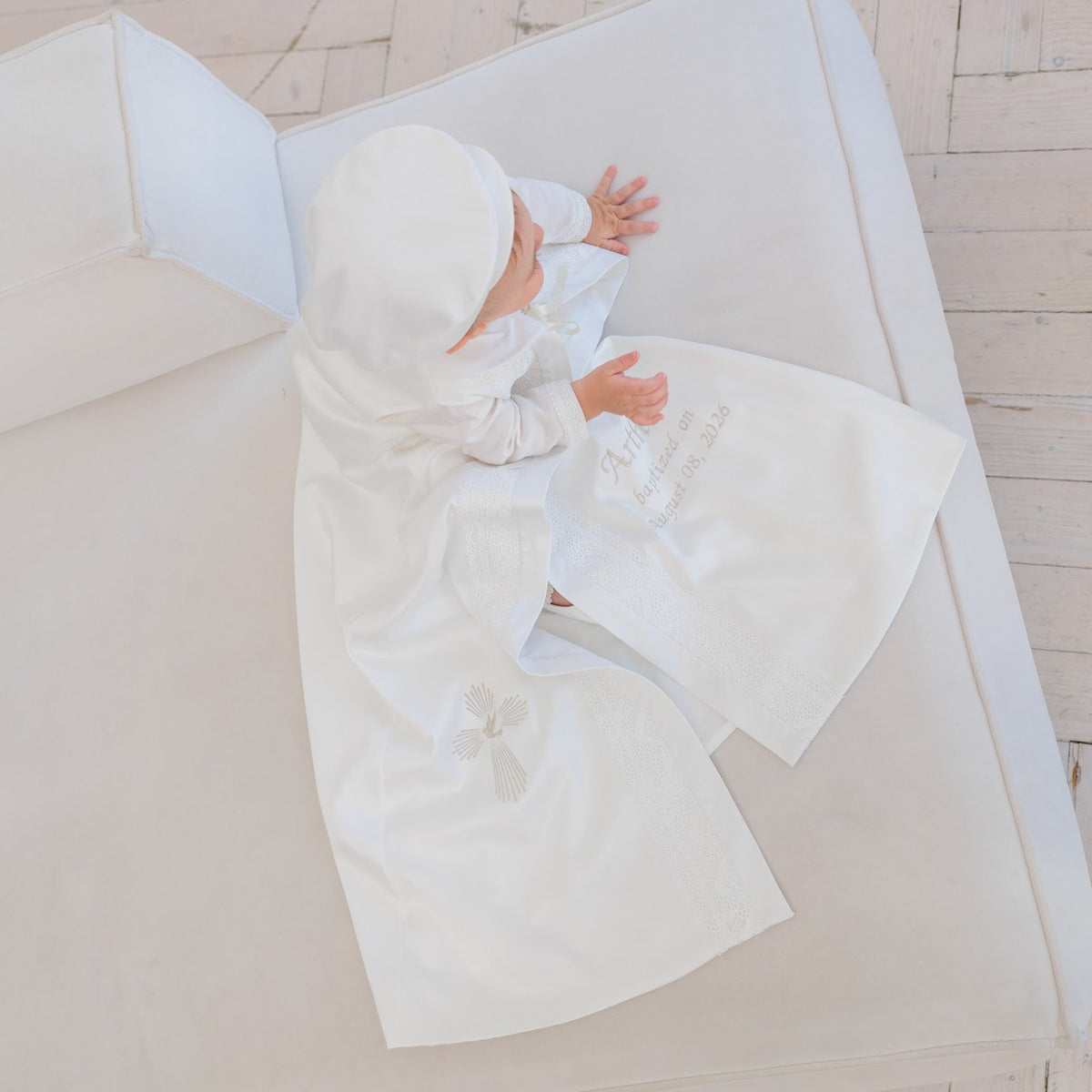 A baby wearing a white satin christening gown and matching bonnet, lying on a white cushioned mat over a light wood herringbone floor.