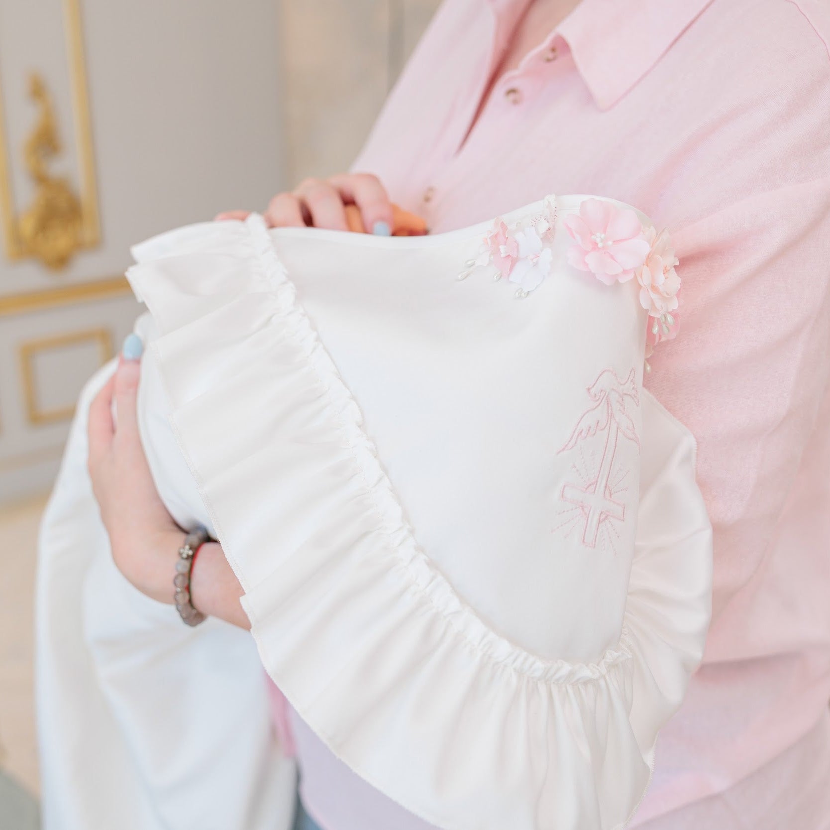 A person in a light pink shirt holds a white ruffled baby blanket or swaddle with pink floral embroidery and ribbon details, set against an elegant interior background with gold-trimmed walls and a pale blue chair.