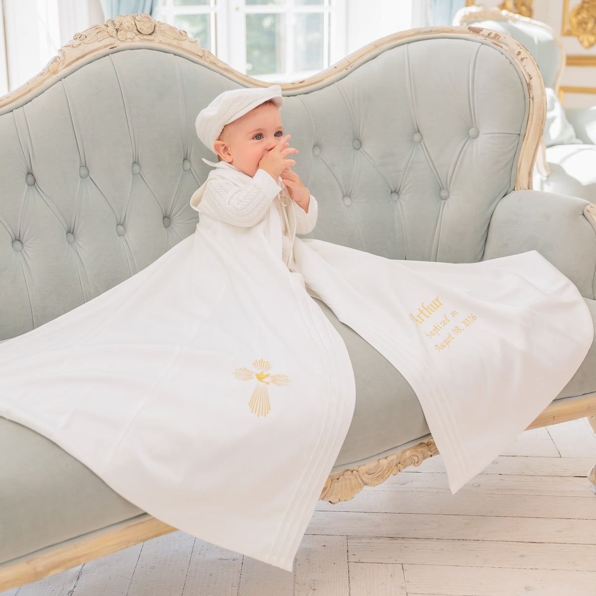A baby wearing a white bonnet and white outfit sits on a tufted pale green sofa, wrapped in a white swaddle blanket with gold embroidered details. The scene is set in a bright, elegant room with light-colored wood floors and windows.