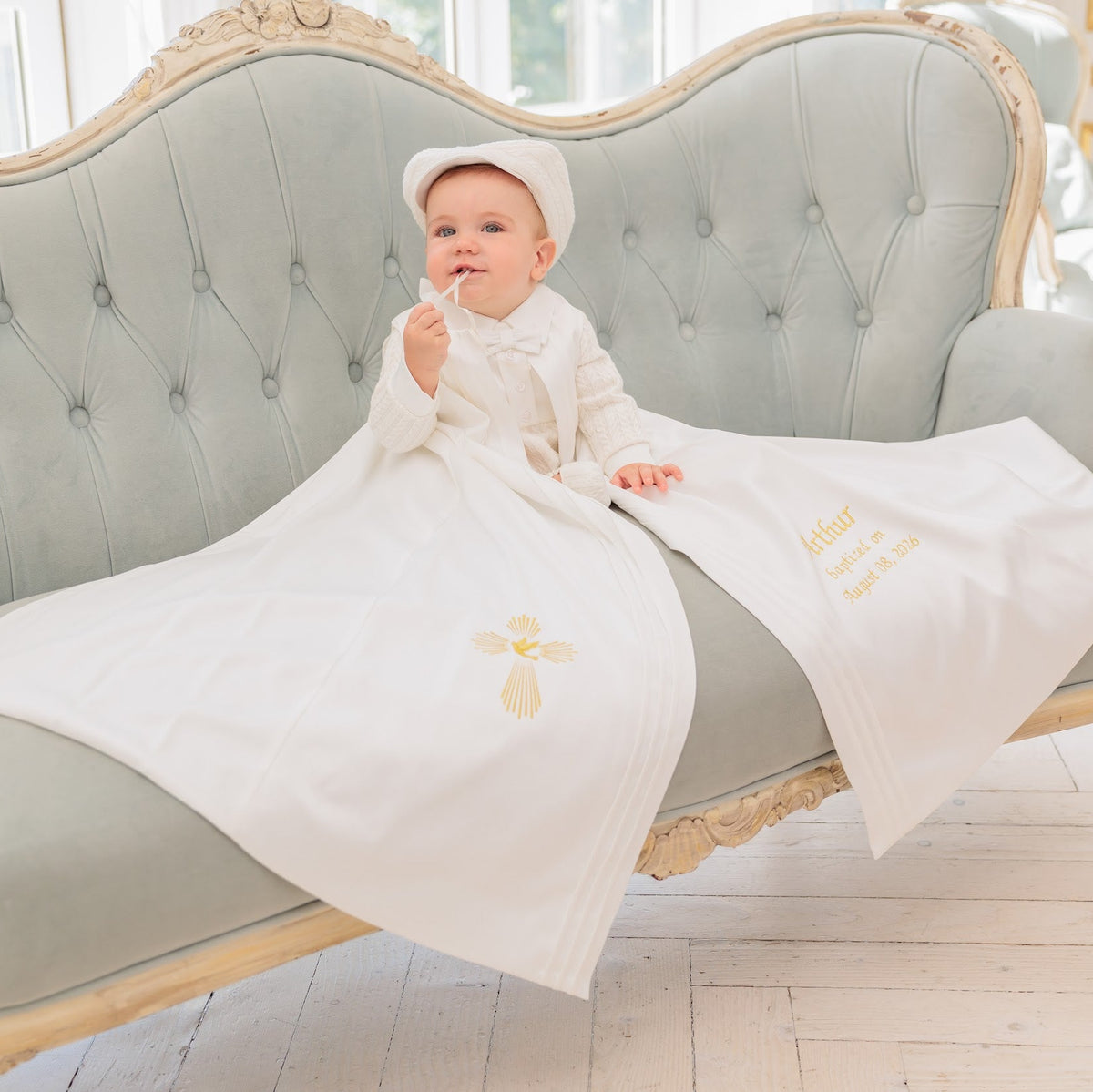 A baby wearing a white christening gown and matching hat sits on a tufted light blue-green sofa, wrapped in a white baptismal blanket with a gold cross and the name 'Isabel' embroidered on it. The room has light wood floors and a classic interior.