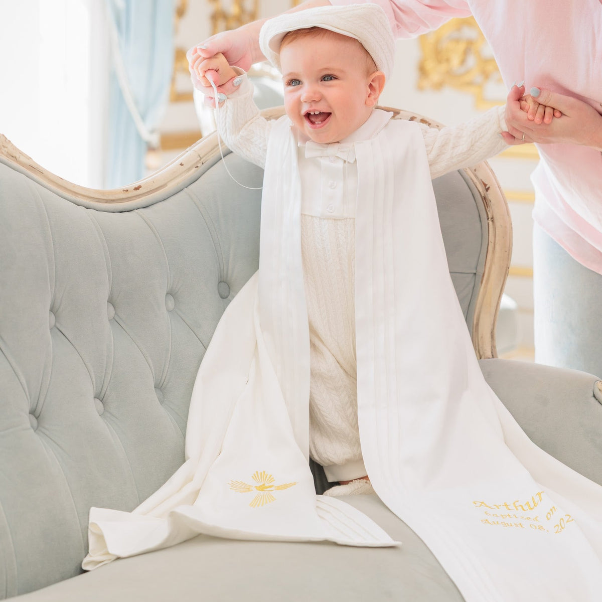 A baby wearing a white traditional outfit and matching hat stands on a tufted gray sofa while holding hands with an adult in a pink top. The outfit has gold embroidered details near the hem.