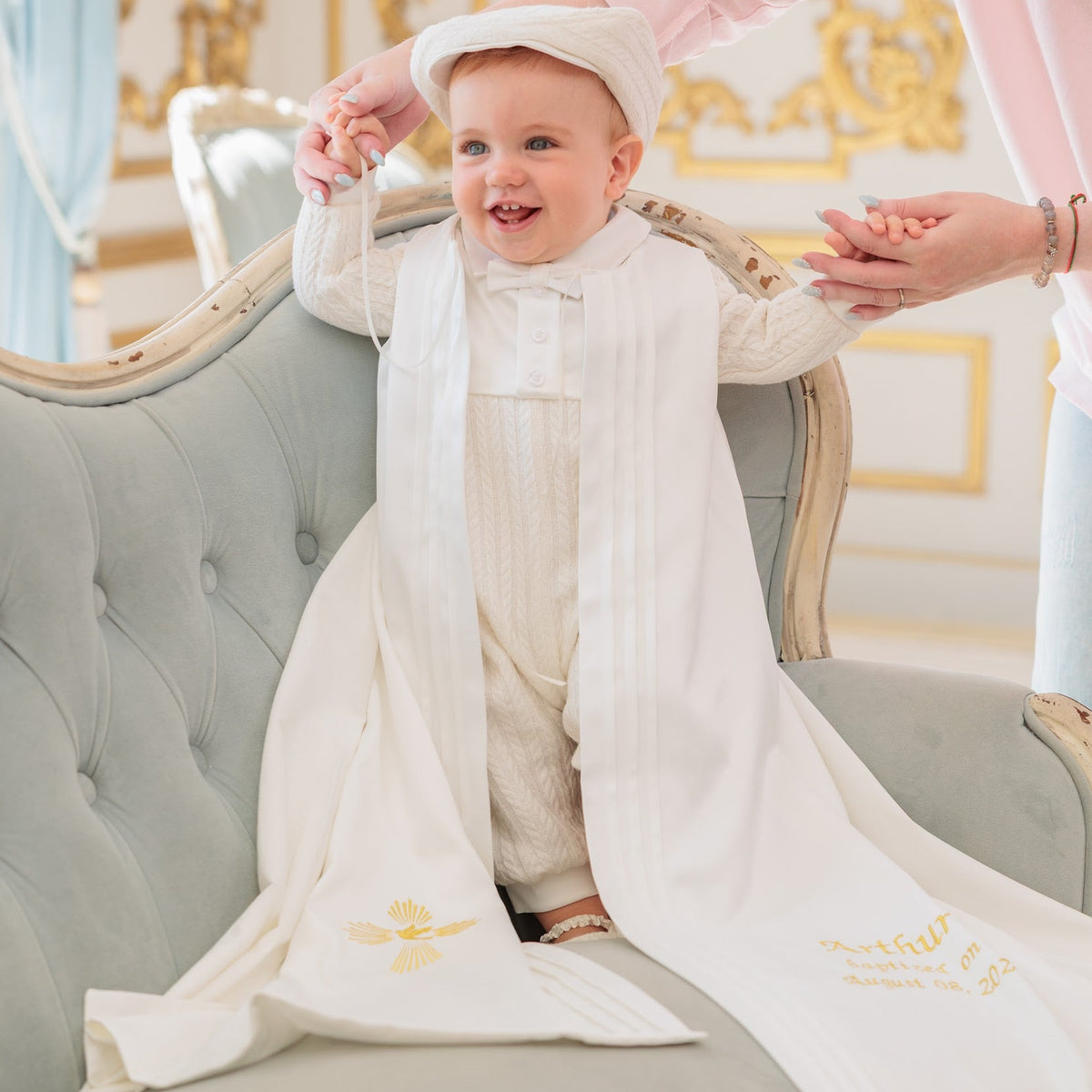 A baby wearing a white traditional boy baptism outfit and matching hat sits on a tufted sofa while an adult in a pink top adjusts the outfit. The scene is set in an elegant room with gold-trimmed walls.