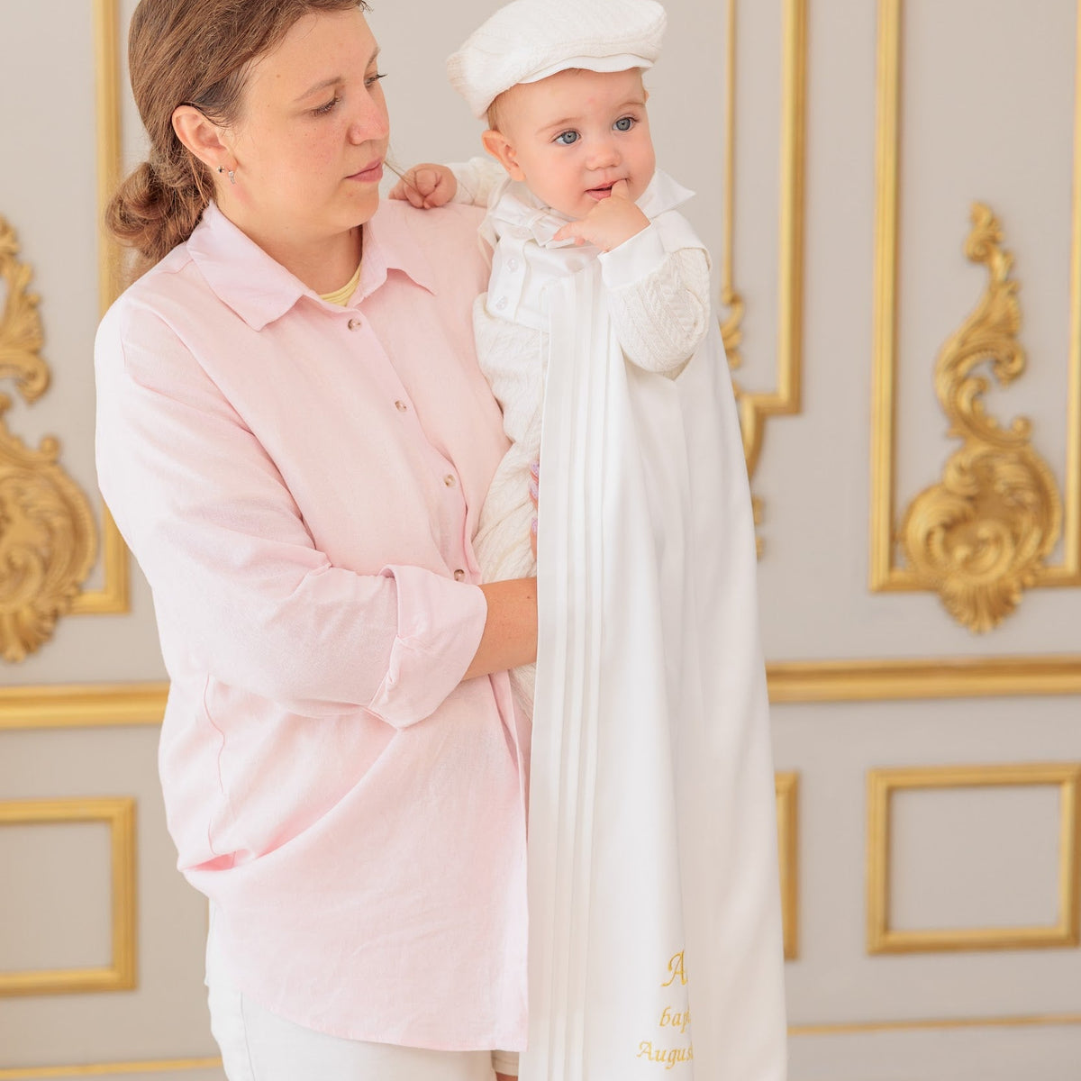 A woman in a light pink shirt and white shorts holds a baby wrapped in a white boy baptism outfit and wearing a white cap, set against an elegant cream and gold paneled wall.