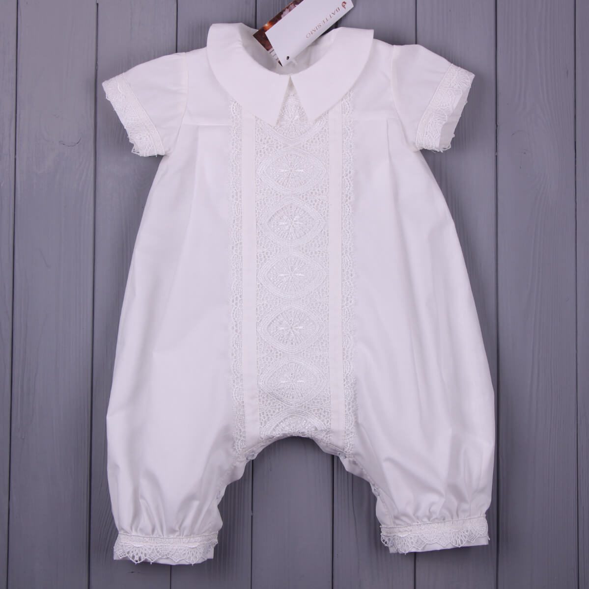 A white baby romper with a Peter Pan collar, short sleeves, lace trim down the front, and lace-cuffed legs, laid flat on a gray wooden plank background.