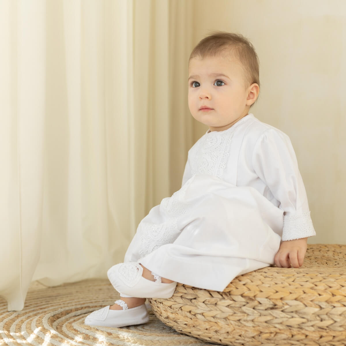 A baby sitting on a woven round ottoman, wearing a white long-sleeve Boy Christening Gown with embroidered detailing and white shoes, set against a soft beige curtain background.