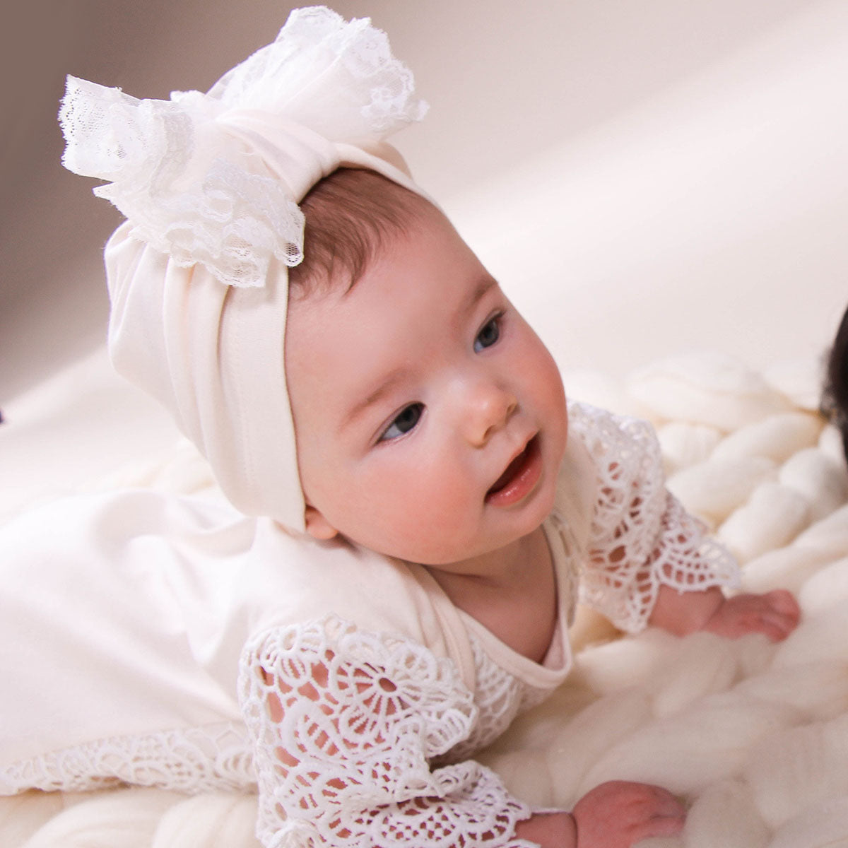 Baby wearing a white lace outfit and headband on a soft, neutral background