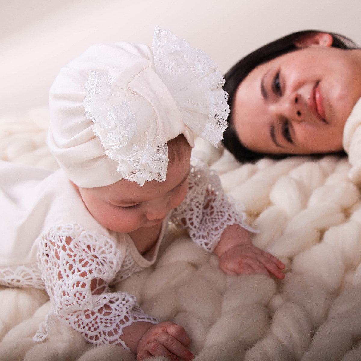 Baby in white lace outfit and bonnet lying next to a woman on a soft white surface