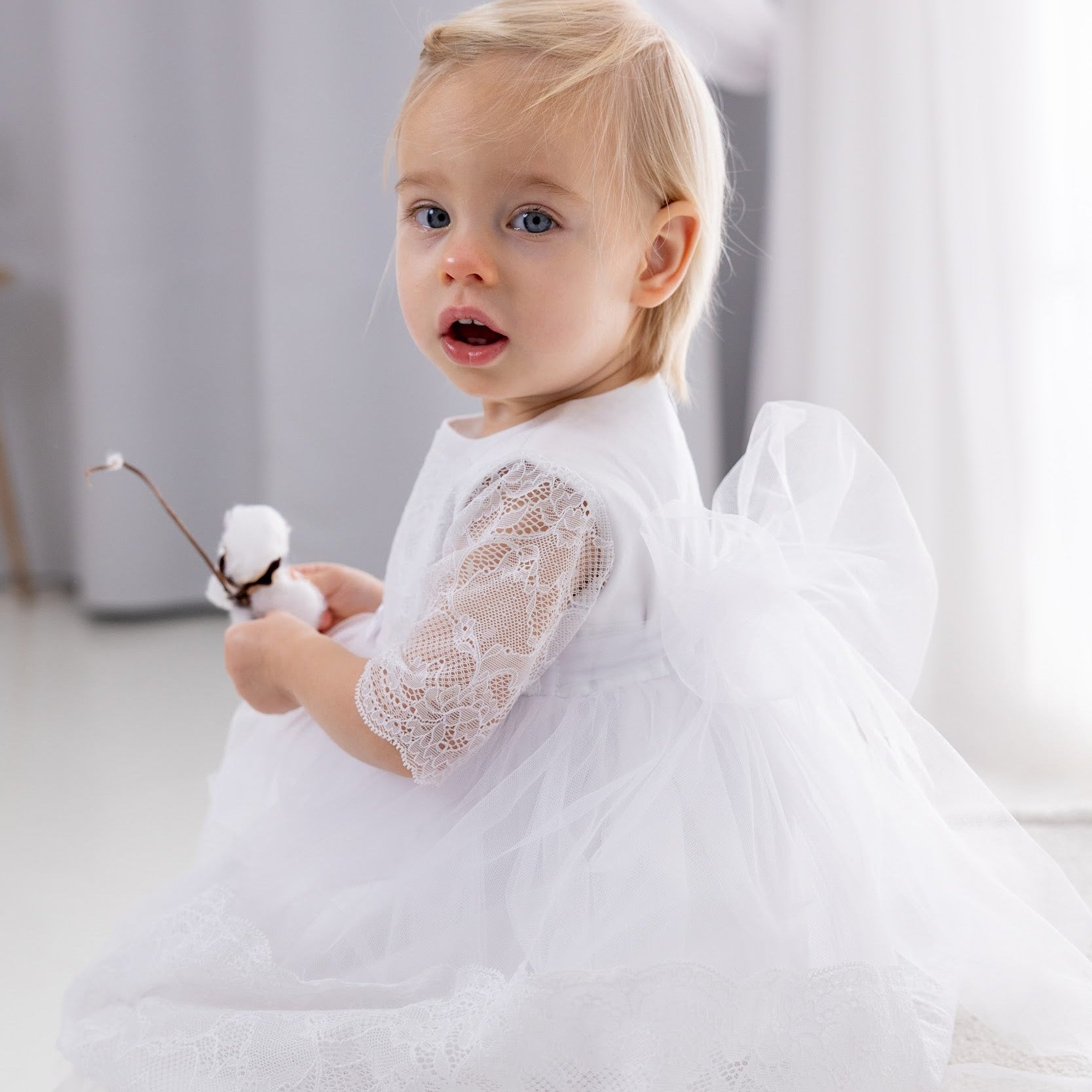 Baby in a white lace baptism dress sitting on a white surface with white curtains in the background