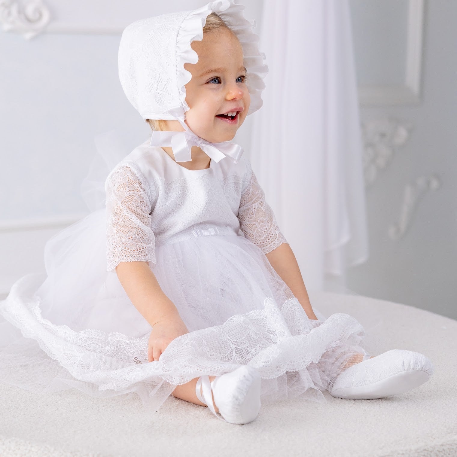 Baby in a white baptism outfit sitting on a white surface with a white background