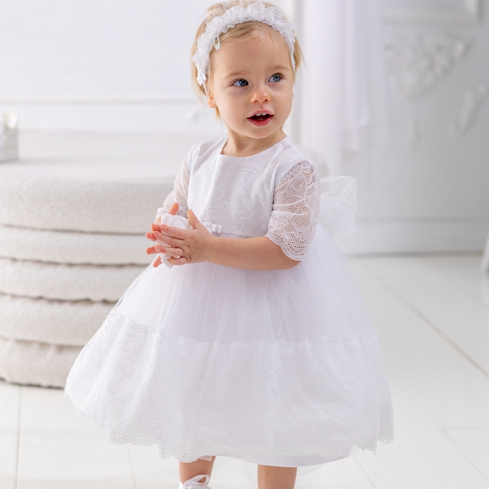 Child in a white dress standing on a white floor with a white curtain in the background
