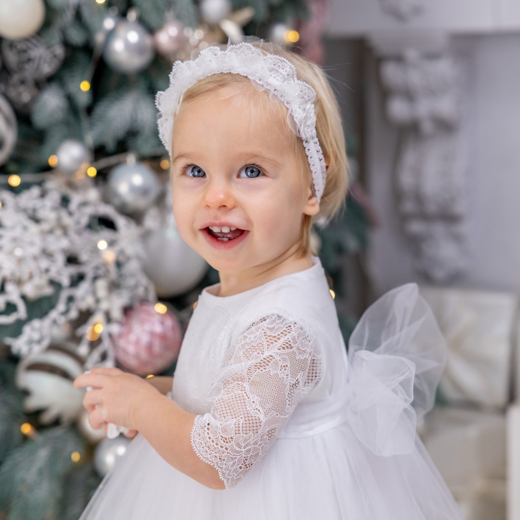 Child in a white lace dress standing in front of a decorated Christmas tree.