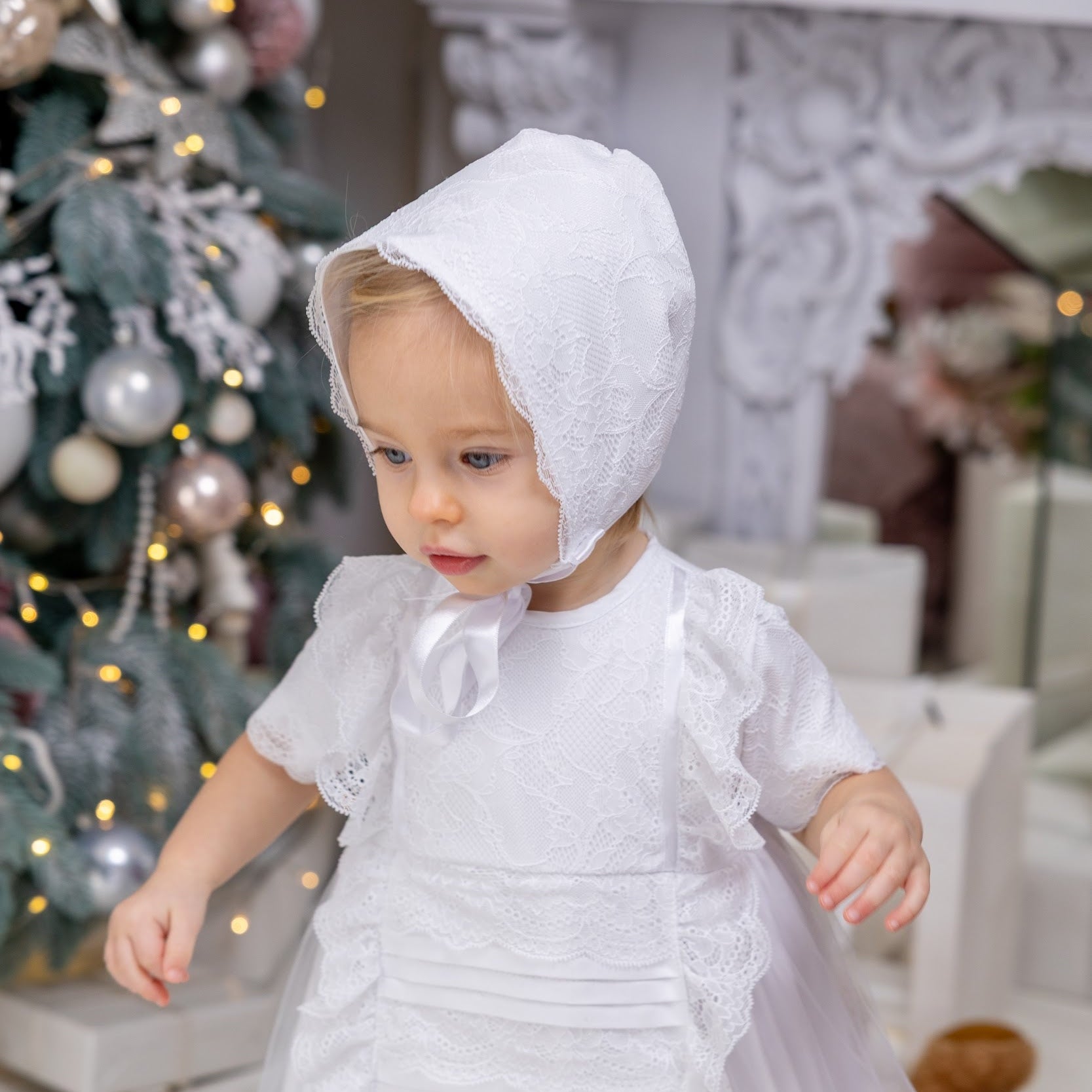 Child in a white dress and bonnet standing in a decorated room with a Christmas tree.