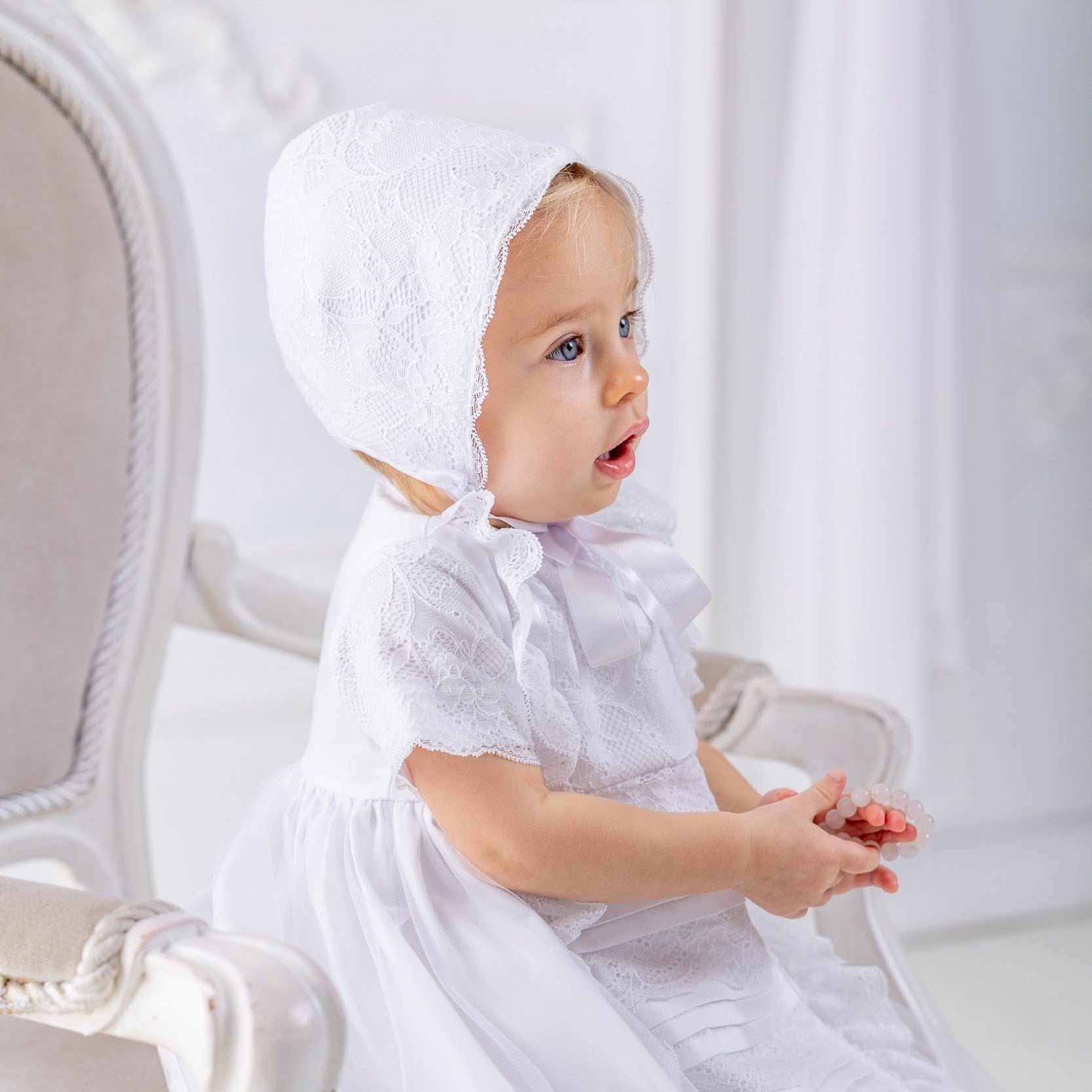 Baby in a white dress and bonnet sitting on a white chair.