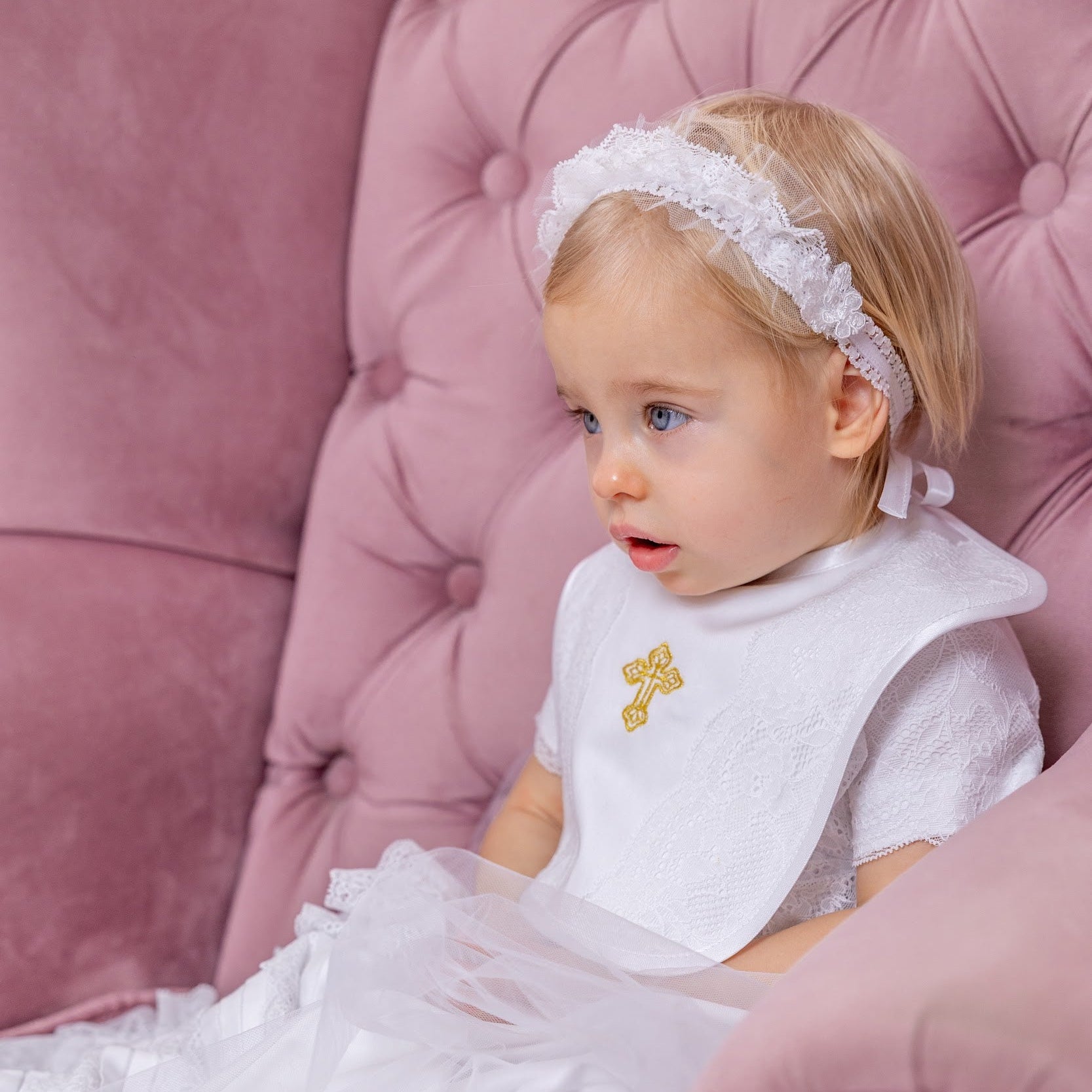 Young child in a white dress with a gold cross sitting on a pink upholstered chair.