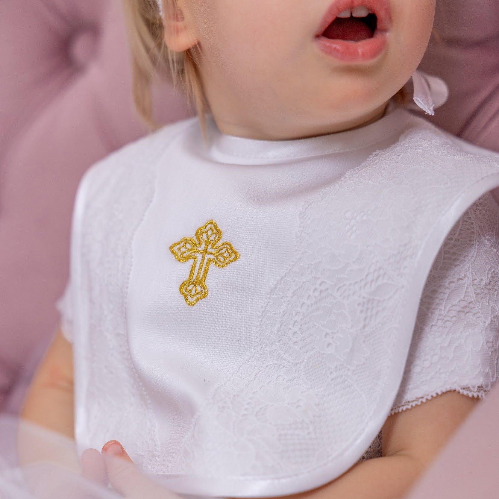 Child wearing a white baptism bib with a gold cross design on a pink background