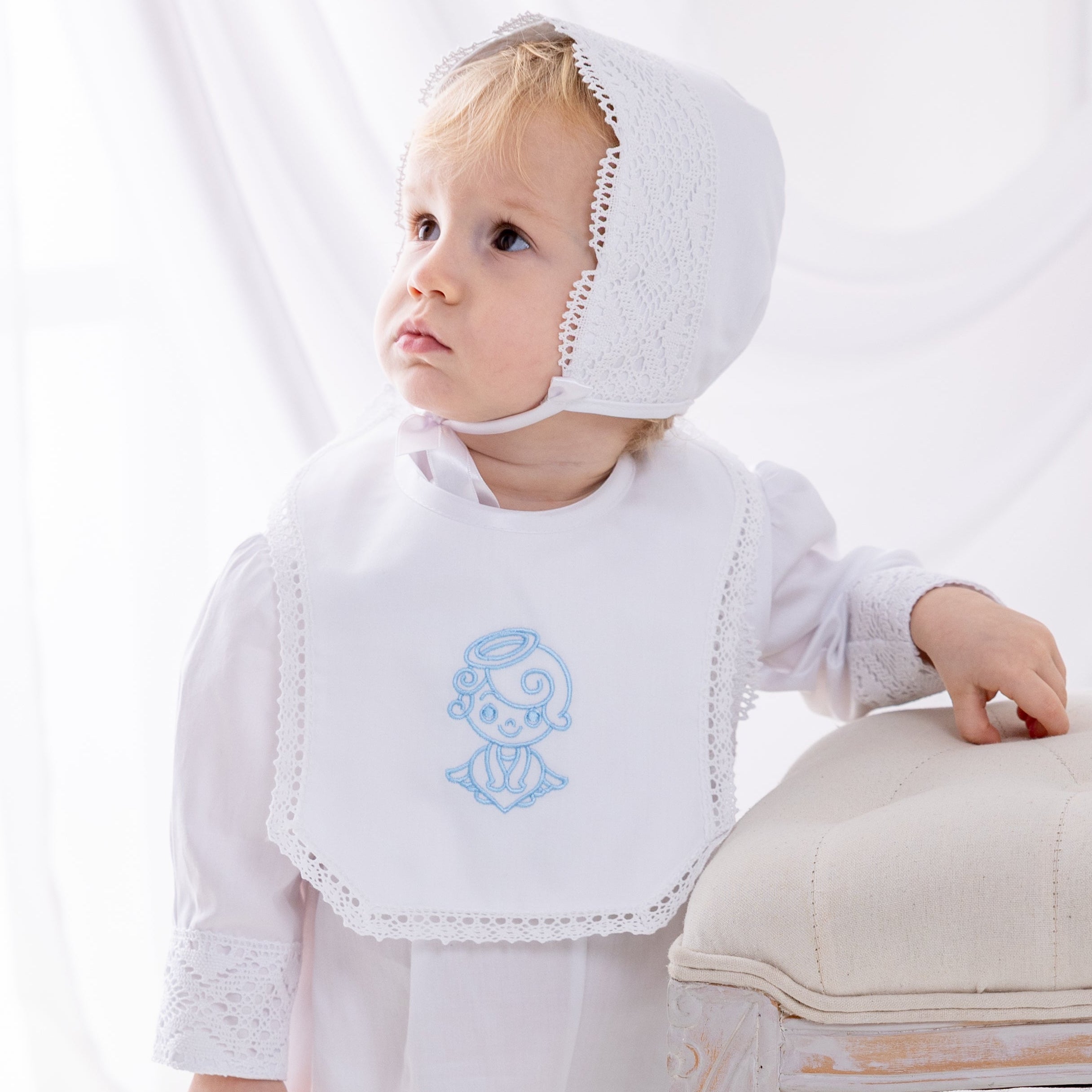 Baby in white outfit with bib and bonnet standing on a white floor.