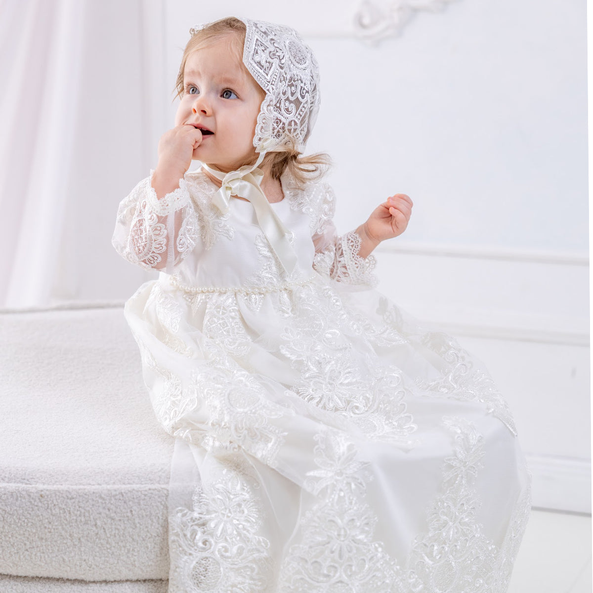Baby in a Long Lace Christening Gown for Girls and bonnet sitting on a white couch.