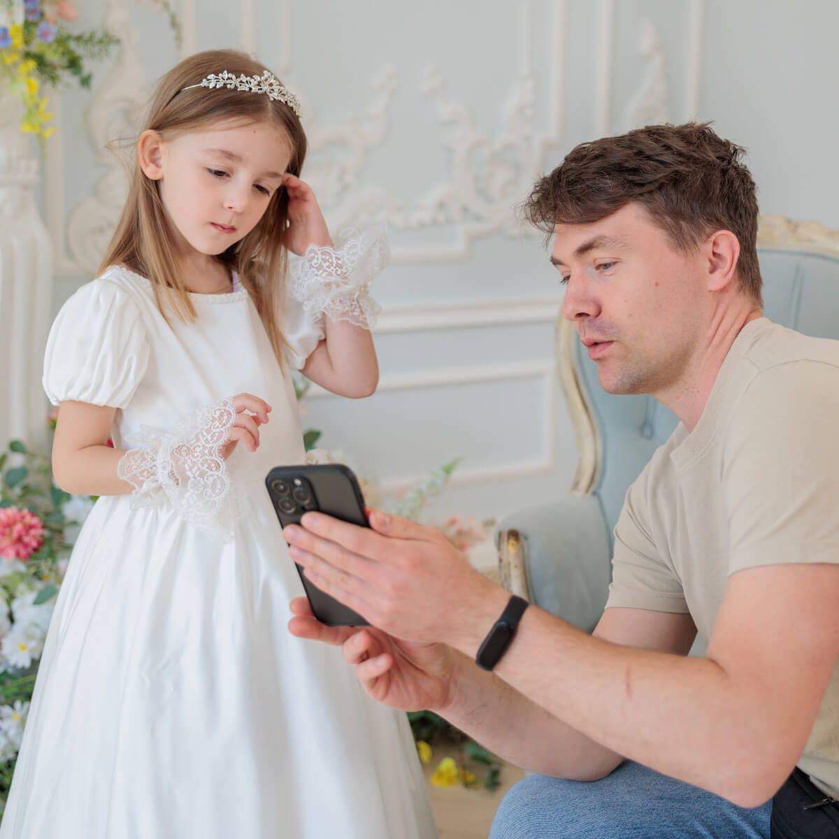Man taking a photo of a young girl in a white dress with a tiara using a smartphone.