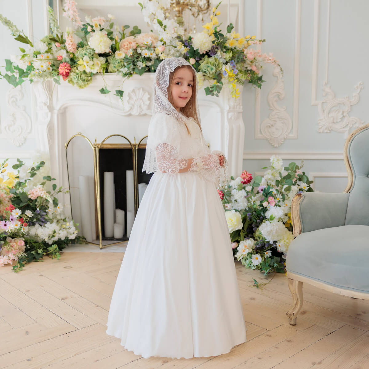 Young girl in  Silk First Communion Dress with Lace Mantilla standing in a decorated room with flowers and a fireplace.