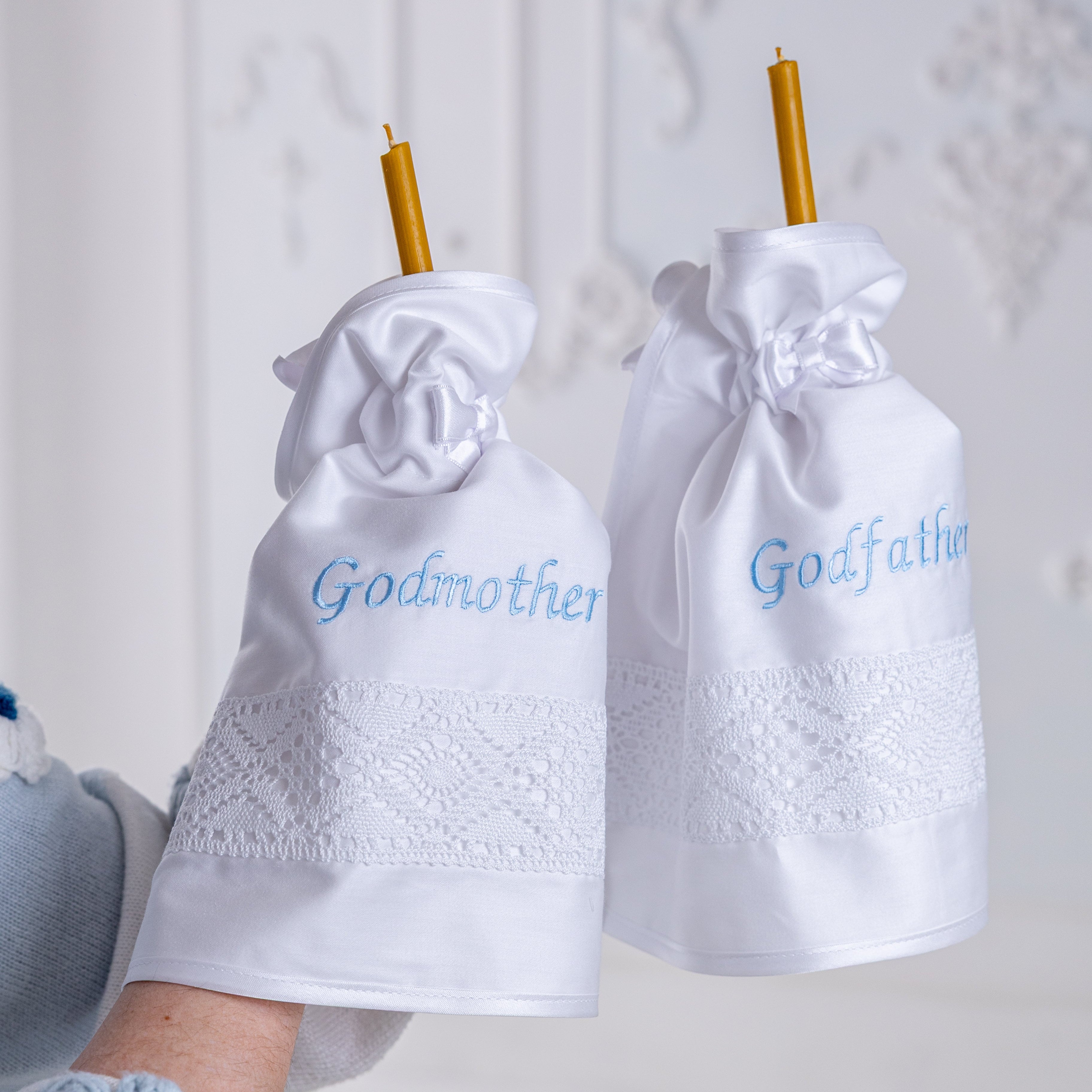 White baptism candle wraps with 'Godmother' and 'Godfather' text, held by a person wearing blue socks.