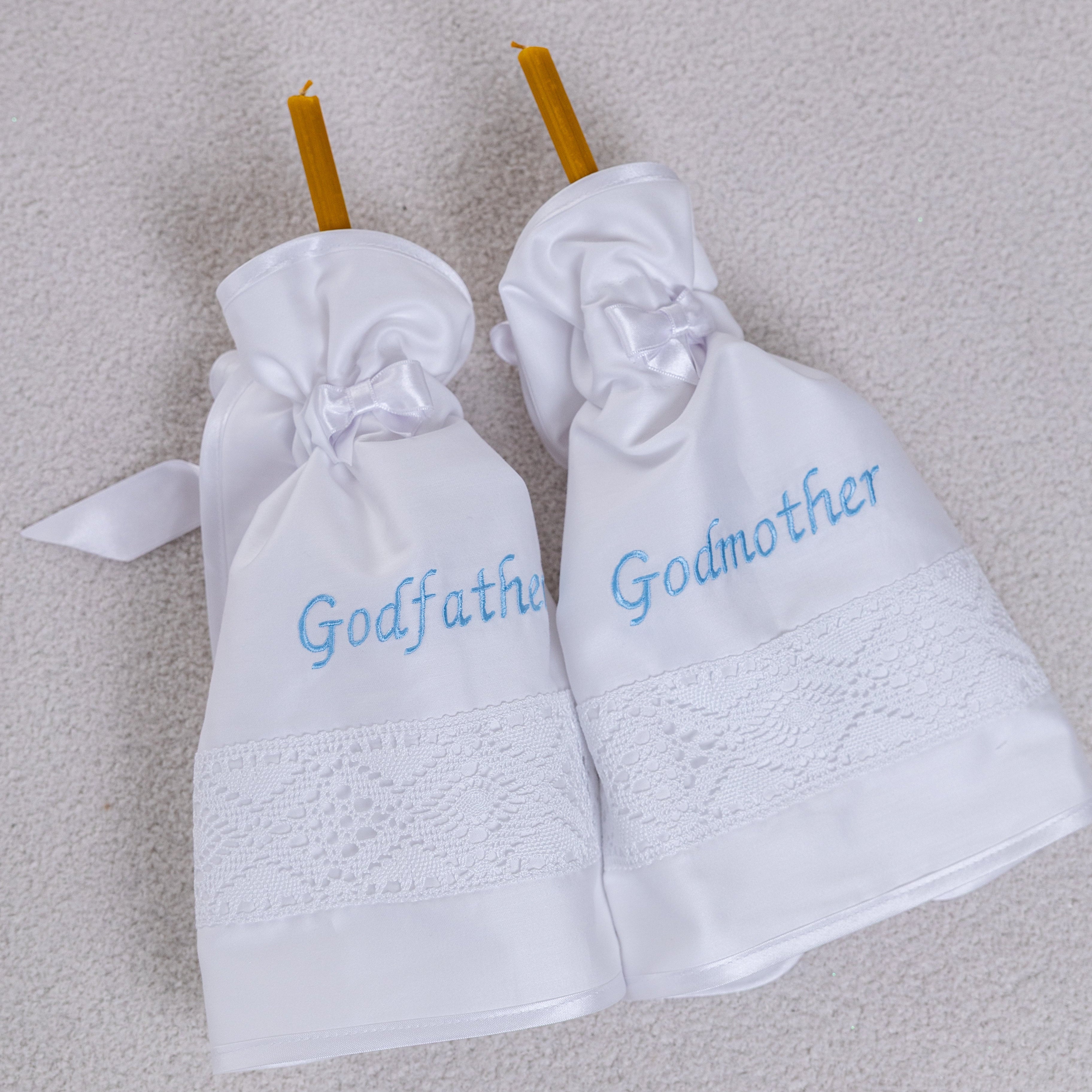 Two white baptism candle wraps labeled 'Godfather' and 'Godmother' on a textured surface.
